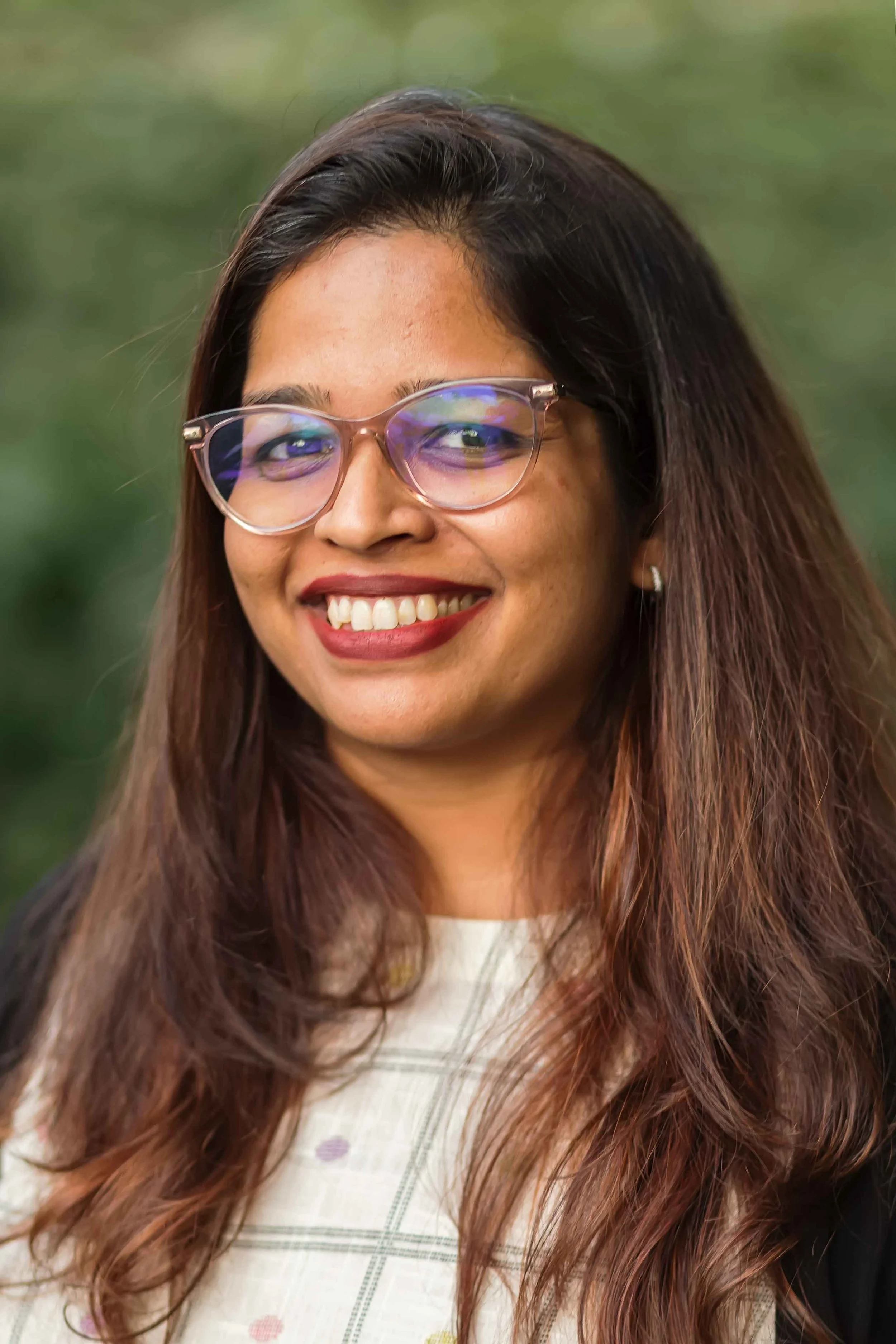 A woman with long brown hair and glasses smiling outdoors with a blurred green background.