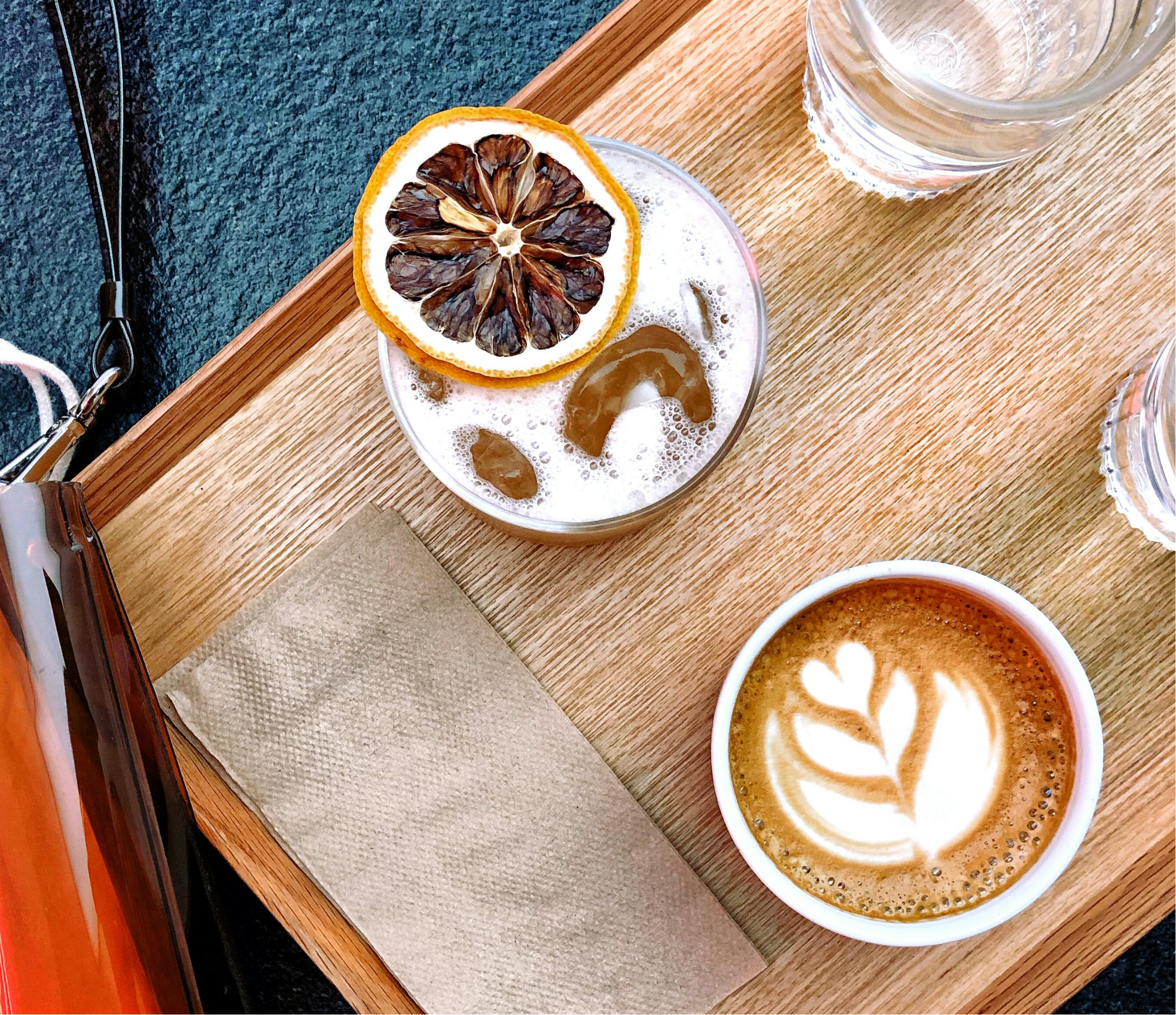 A glass of latte with foam art, topped with a dried lemon slice, placed on a wooden tray beside a glass of water.
