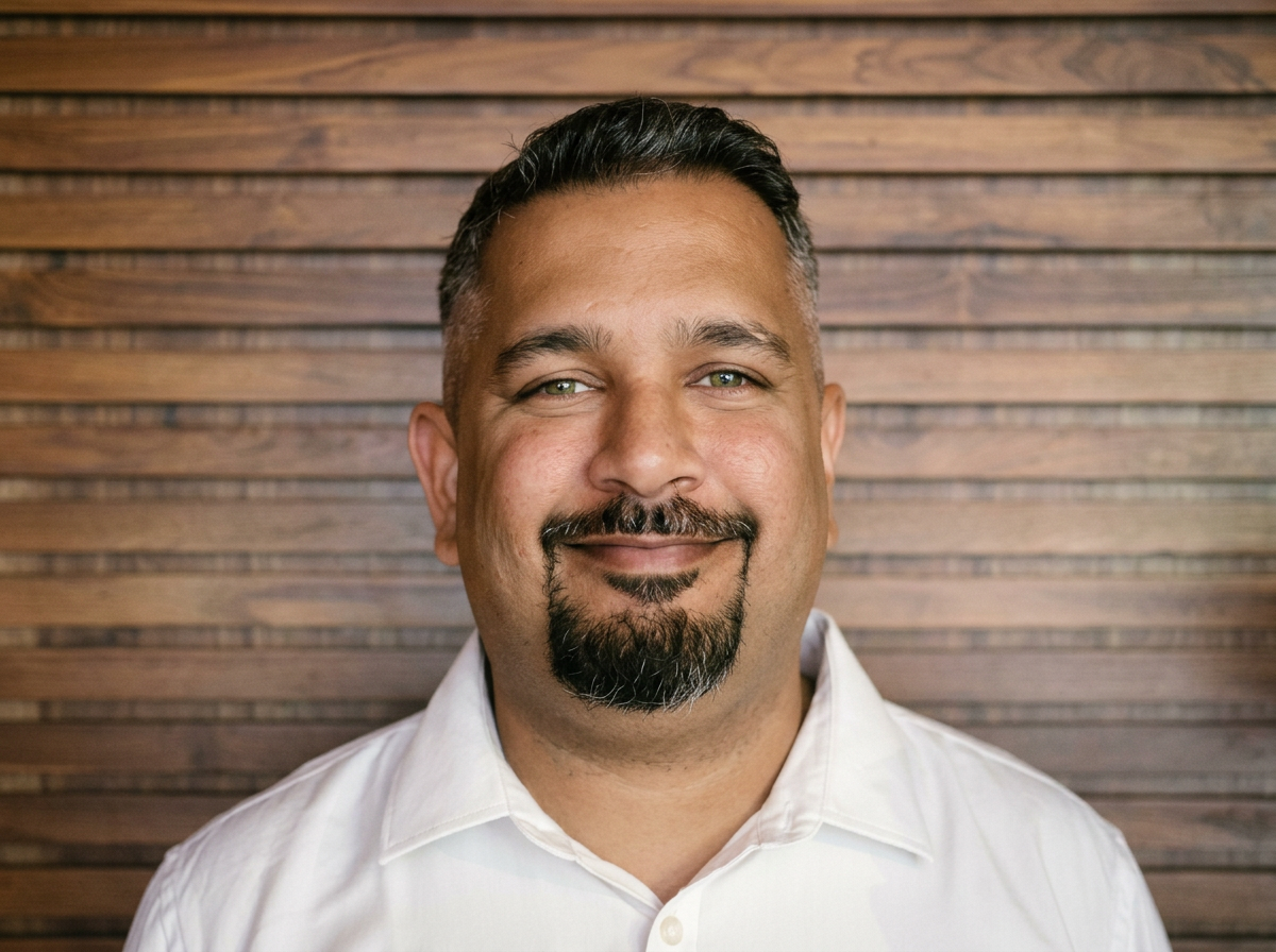 Portrait of a smiling man with short dark hair, a goatee, and a mustache, wearing a white collared shirt, standing in front of a wooden wall.