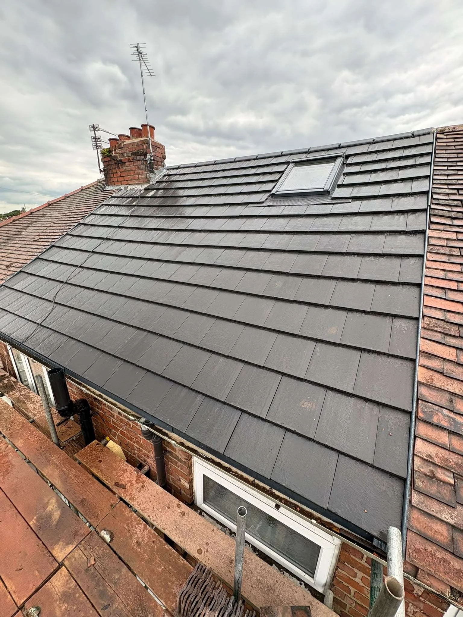 A rooftop with new dark gray tiles, a skylight window, a brick chimney with multiple TV antennas, and a brick wall with a window below the roofline. There are scaffolding, scaffolding pipes, a broom, and a rust-red ledge in the foreground.