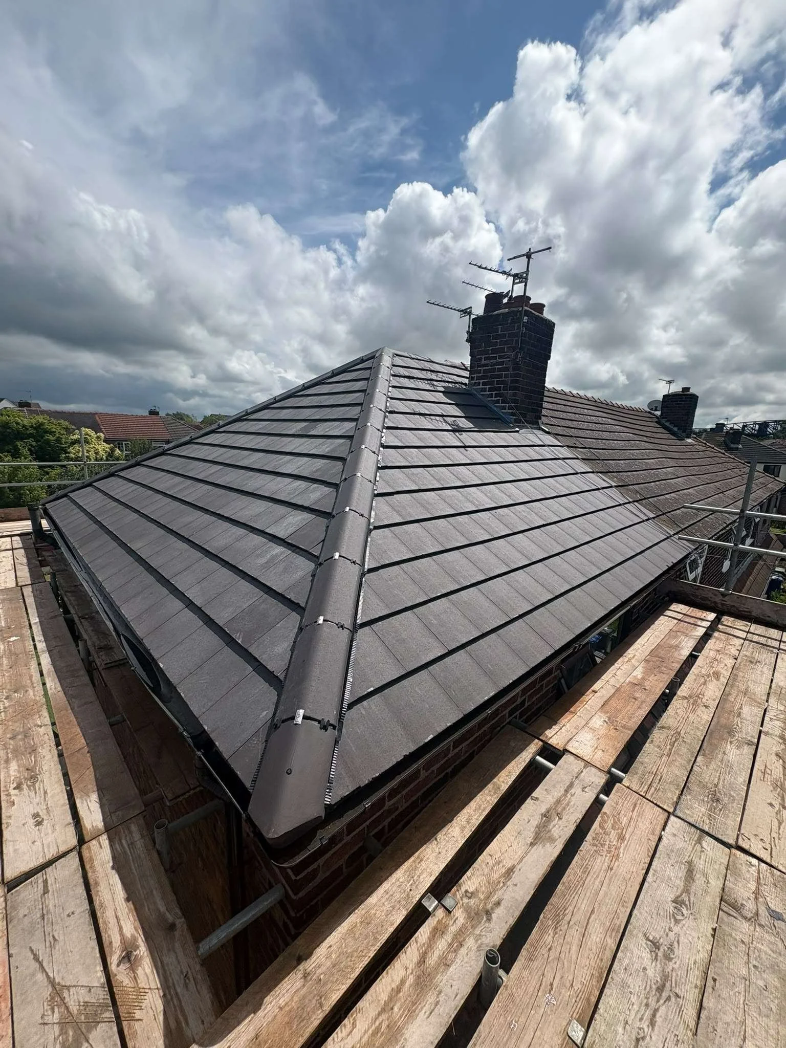 View of a rooftop with a chimney and television antennas, under a partly cloudy sky, with surrounding rooftops and trees.