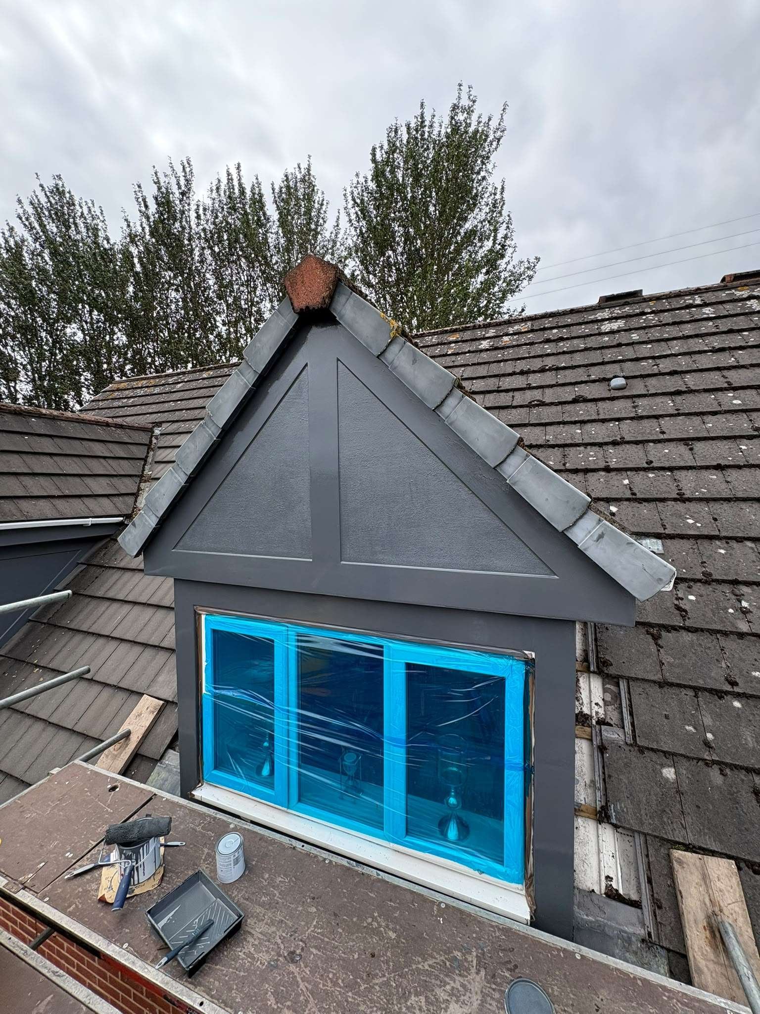 View of a house roof with a dormer window under renovation, construction tools and supplies on a work table, overcast sky, and trees in the background.