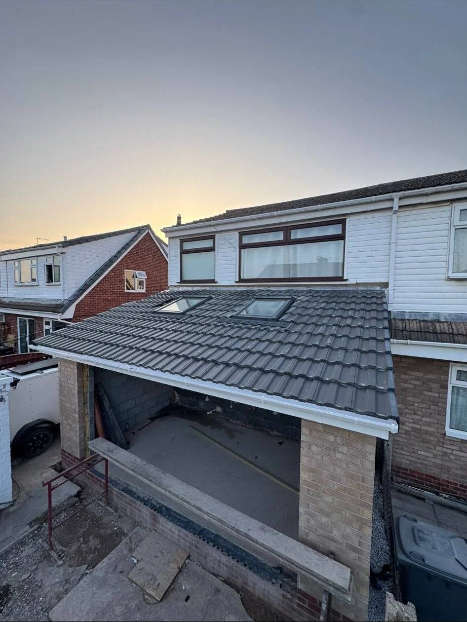 View of a residential house under construction with a tiled roof, skylights, and an open garage area during dusk, with neighboring houses in the background.