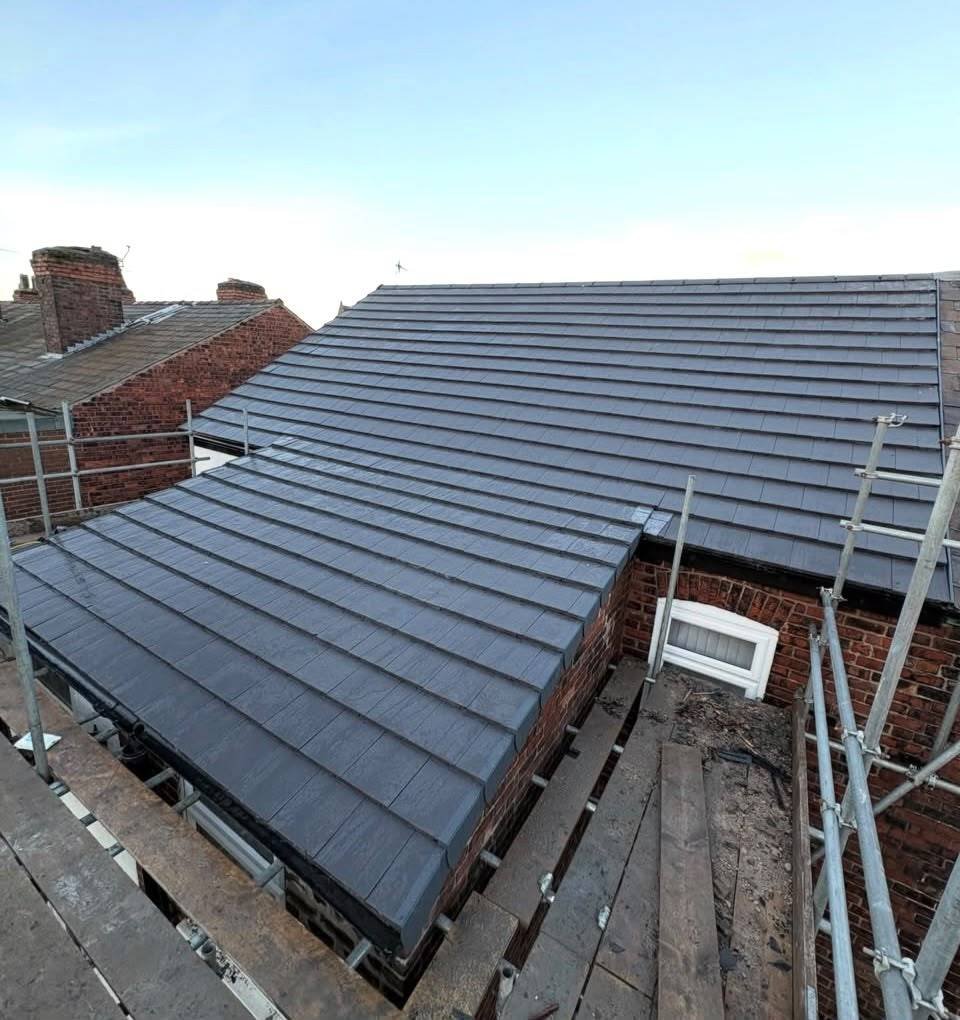 View of a newly installed gray tiled roof on a brick house, with scaffolding surrounding it and a small window near the roof's edge.