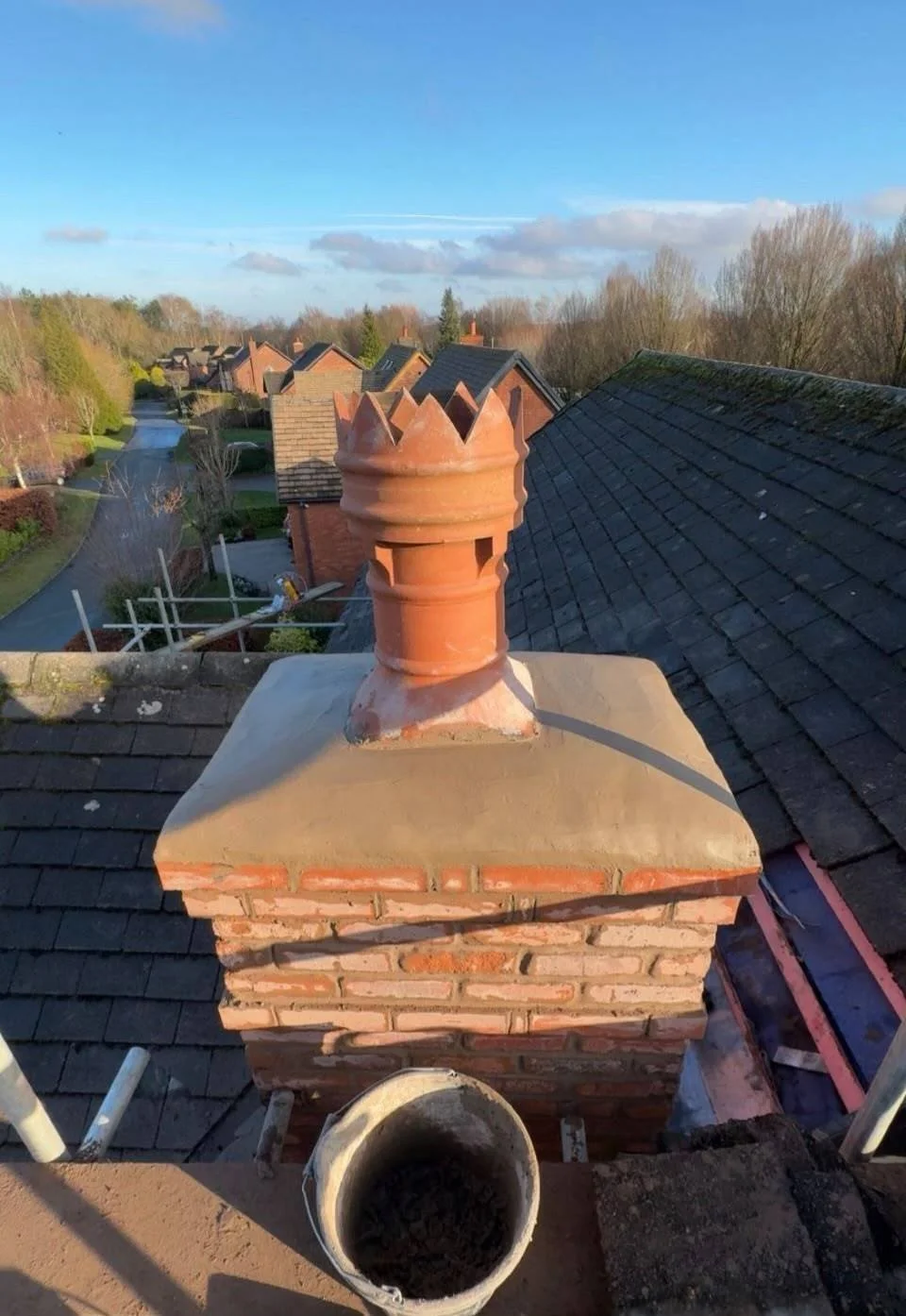 A brick chimney with a brick crown cap on a rooftop, with a blue sky and neighborhood houses and trees in the background.