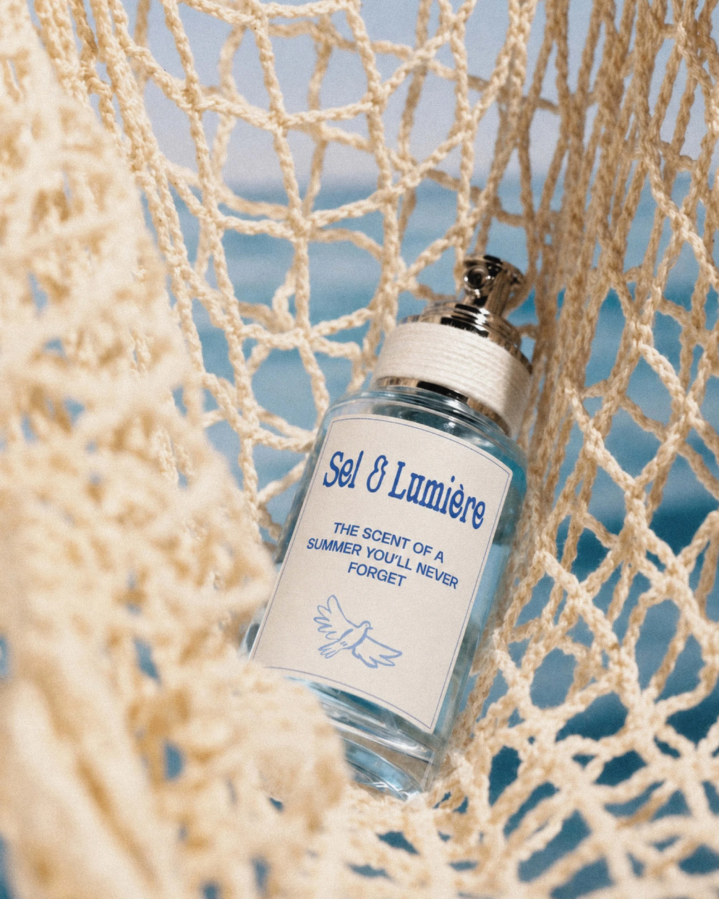 Une bouteille de parfum posée dans un filet de pêche en corde de couleur beige, avec une étiquette blanche et du texte en français, sur fond de ciel bleu et d'eau, évoquant une atmosphère estivale et marine.
