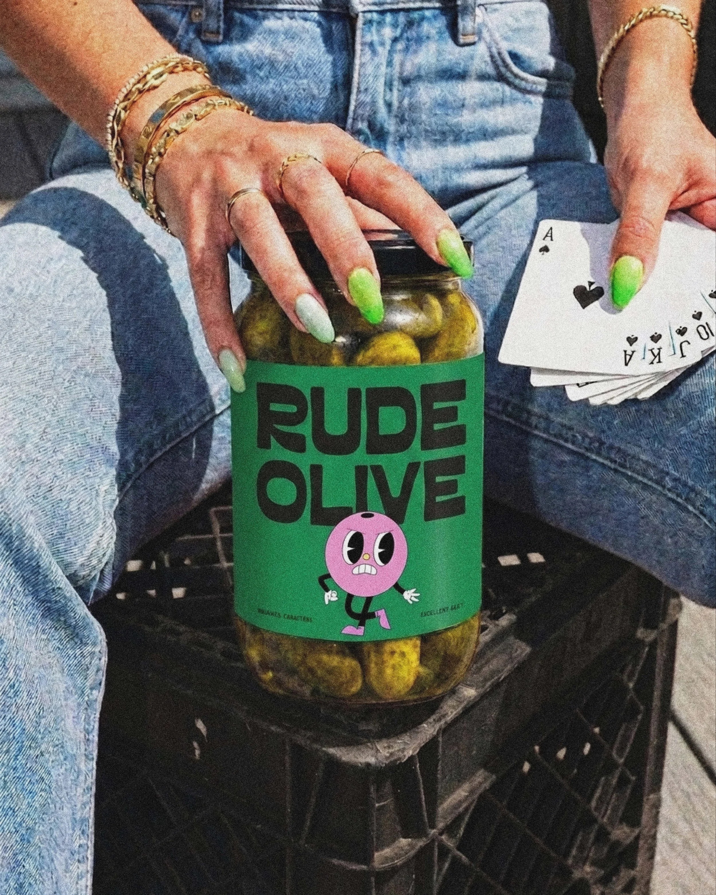 Une main avec des ongles vert fluo tient un pot de cornichons. La personne porte des bijoux dorés au poignet et des bagues. Elle tient également une paire de cartes, dont une est une as de pique.