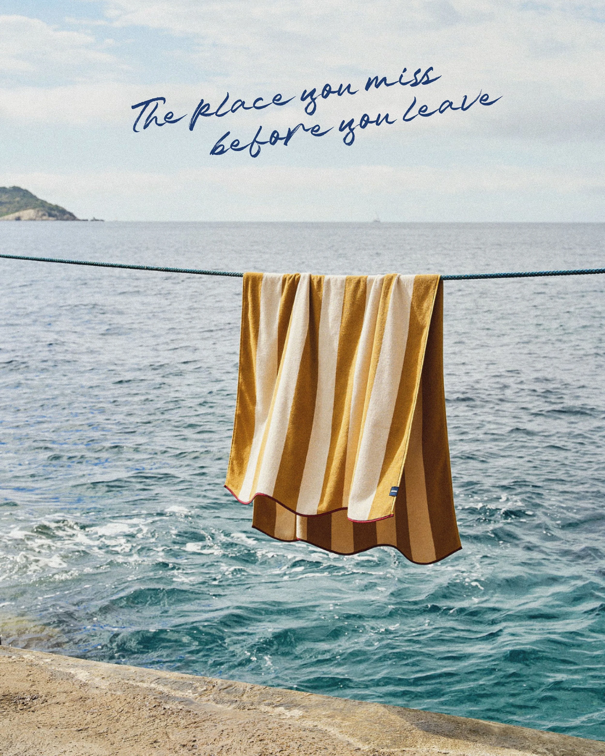 Une corde tendue au-dessus de la mer avec un vêtement suspendu. En arrière-plan, l'océan avec une petite île ou une péninsule à gauche et un ciel partiellement nuageux. Texte en surimpression disant 'The place you miss before you leave'