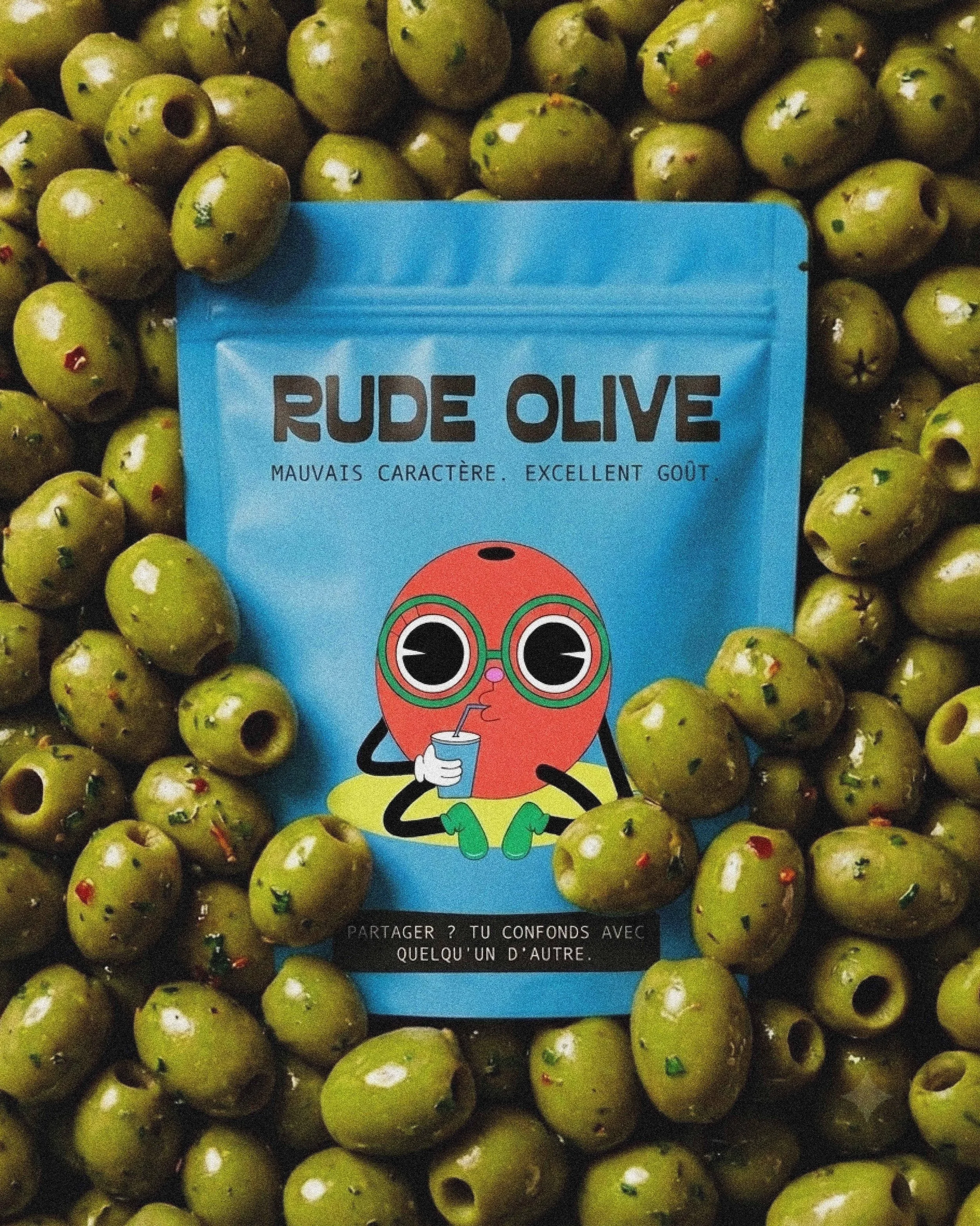 Un panier rempli d'olives vertes avec un emballage bleu au centre. L'emballage porte l'inscription « RUDE OLIVE » et une illustration d'une figure cartoon jeune qui boit dans un gobelet avec une pailles.
