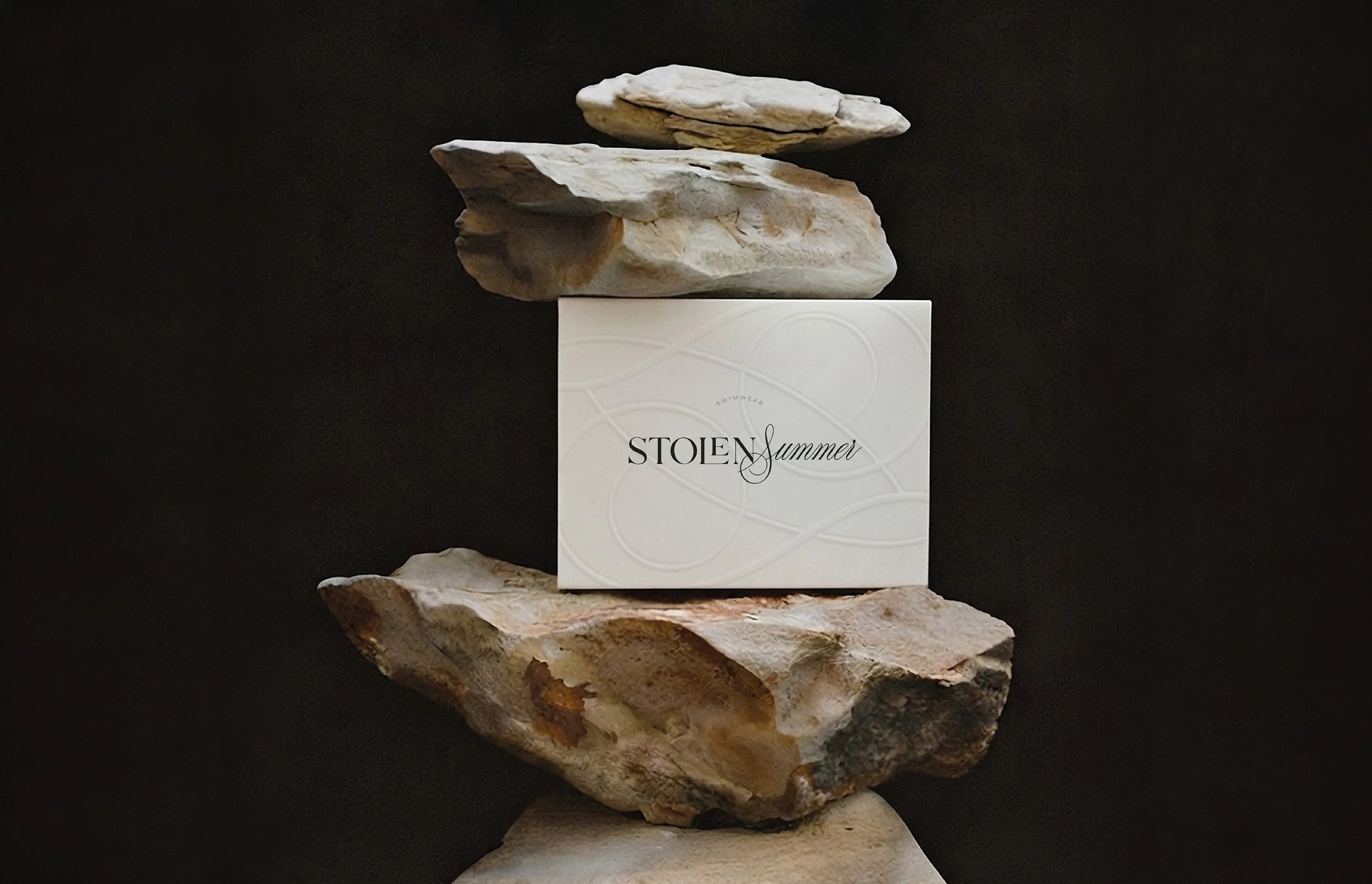 Une composition artistique avec des rochers empilés, surmontée d'une boîte blanche portant l'inscription "STOEN summer" et le mot "SWIMWEAR".