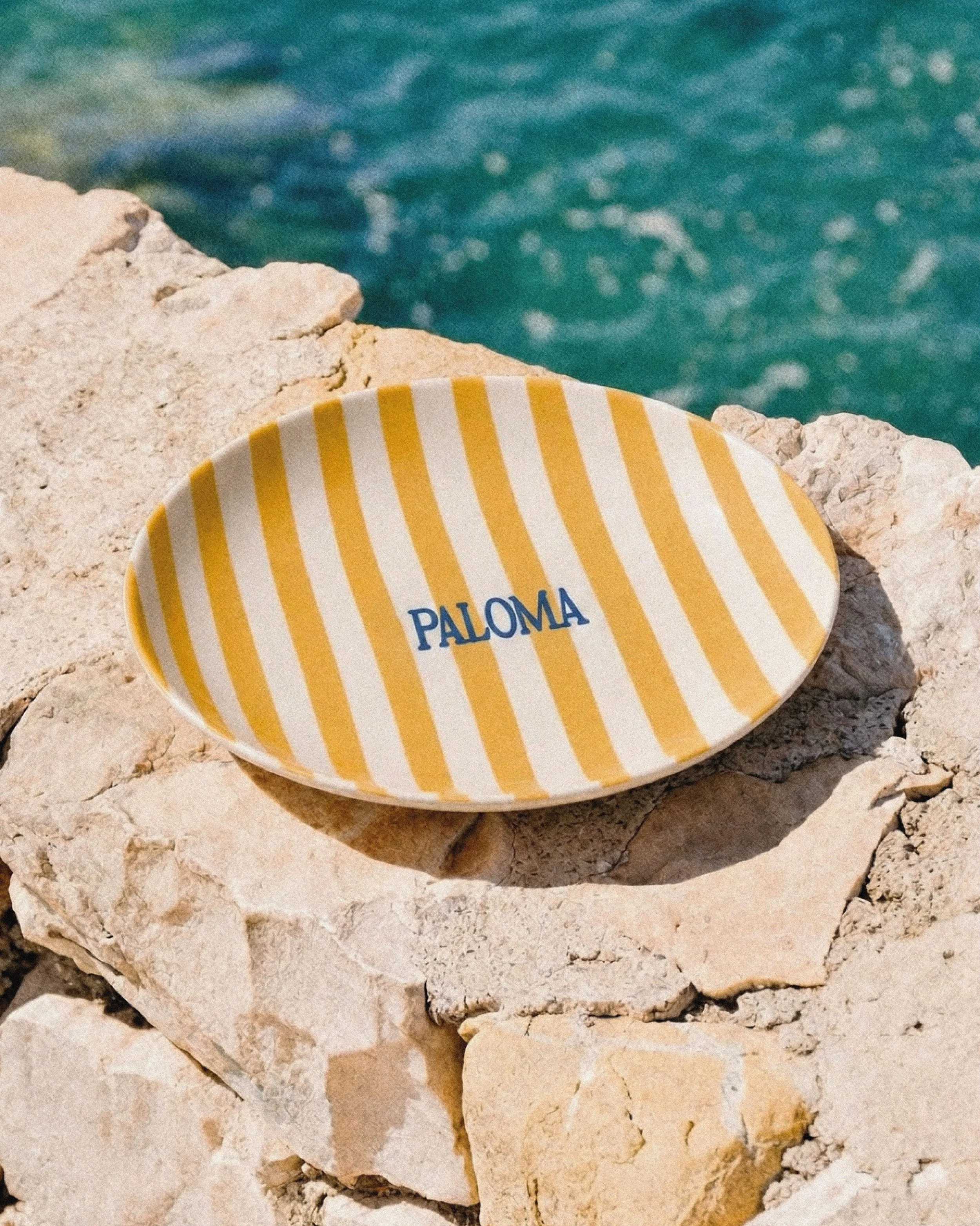 Assiette de taille moyenne avec rayures jaunes et blanches sur fond blanc, posée sur des rochers de pierre claire, avec mer turquoise visible en arrière-plan.