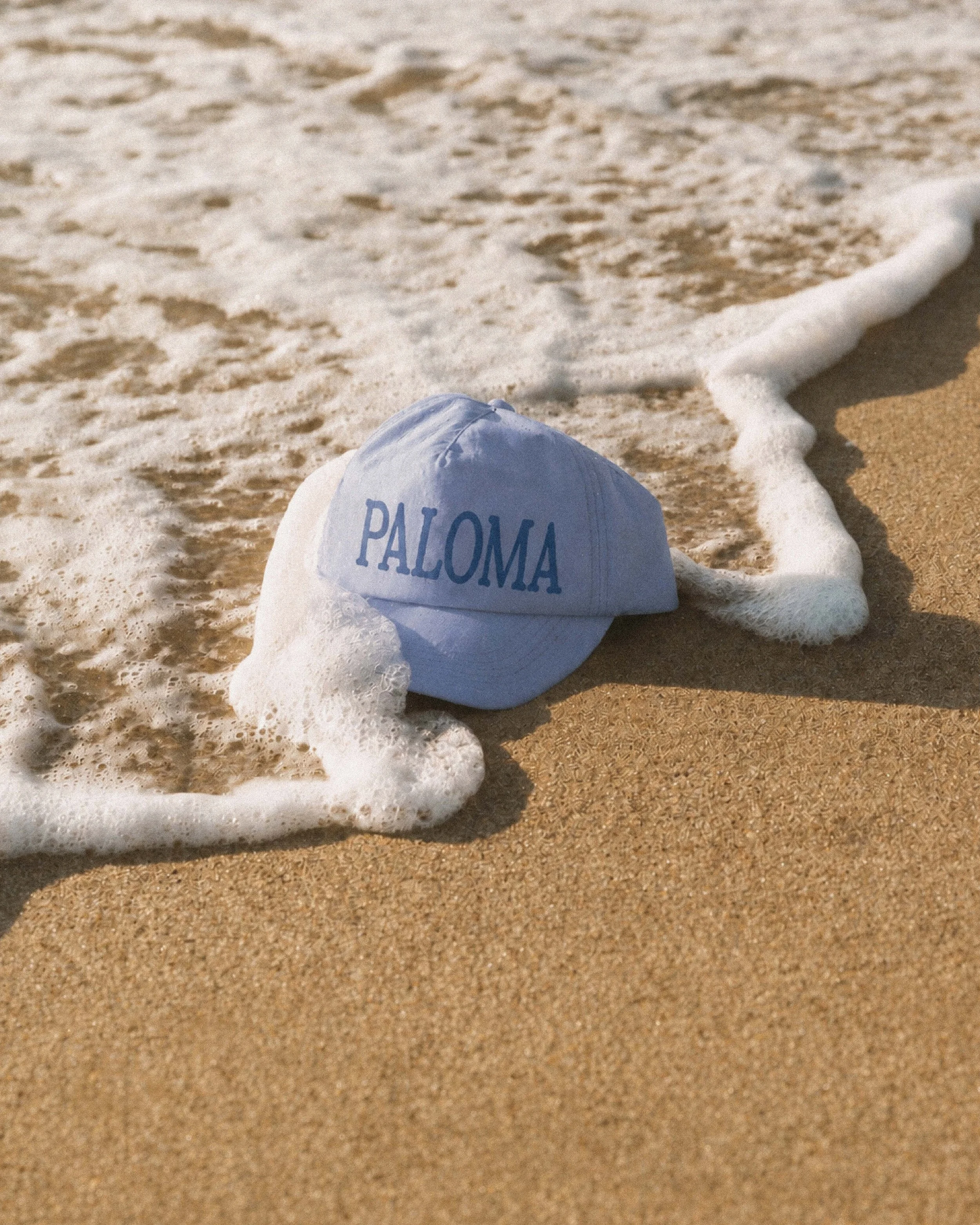 Un coquillage blanc avec l'inscription 'PALOMA' écrit en bleu, posé sur le sable avec des vagues de mer approchant.