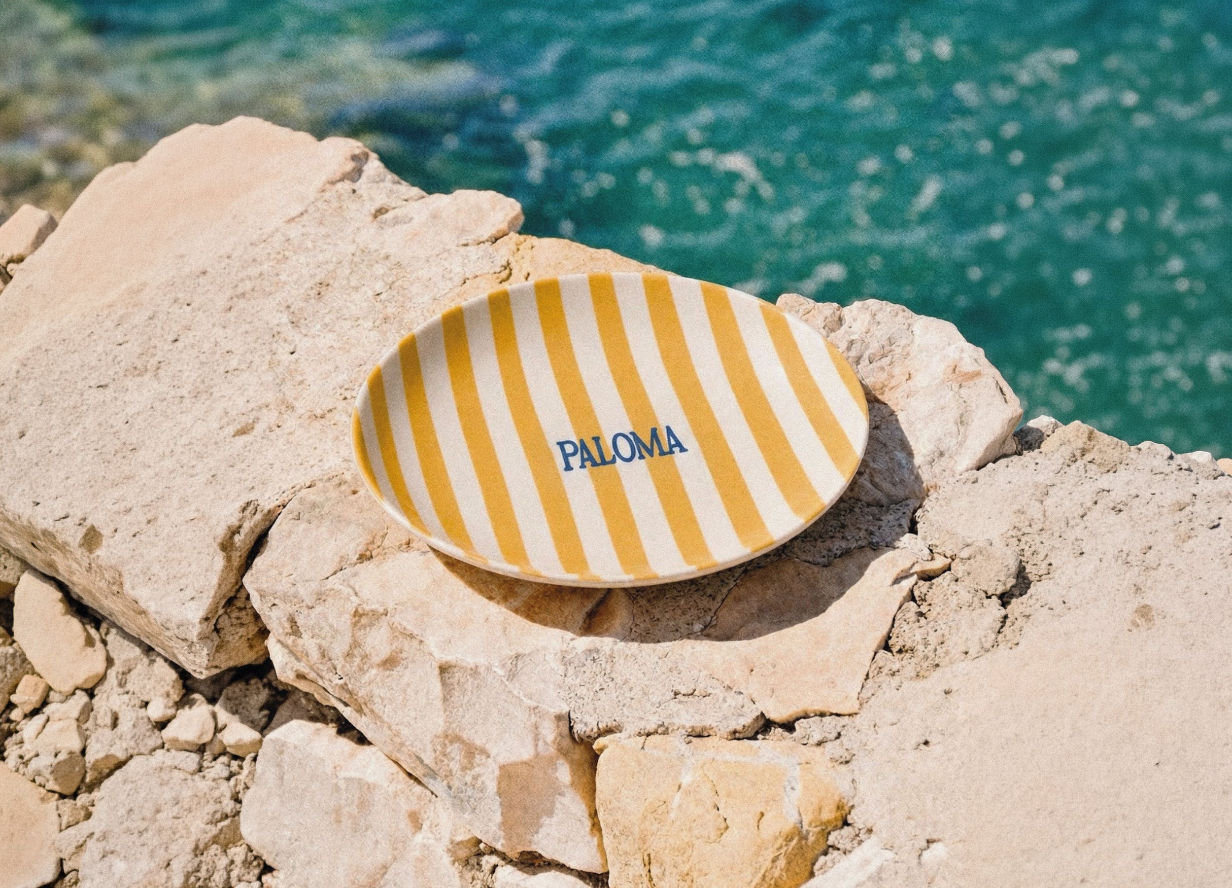 Une assiette jaune et blanche rayée avec le mot "PALOMA" posé sur des rochers de pierre au bord de la mer.