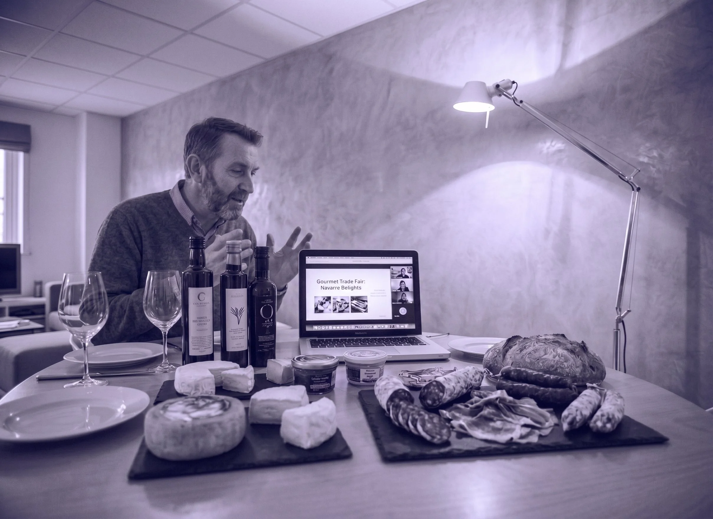 Mesa con queso, embutidos, pan, y vino. Persona en una videollamada en una laptop en un ambiente de oficina o comedor.