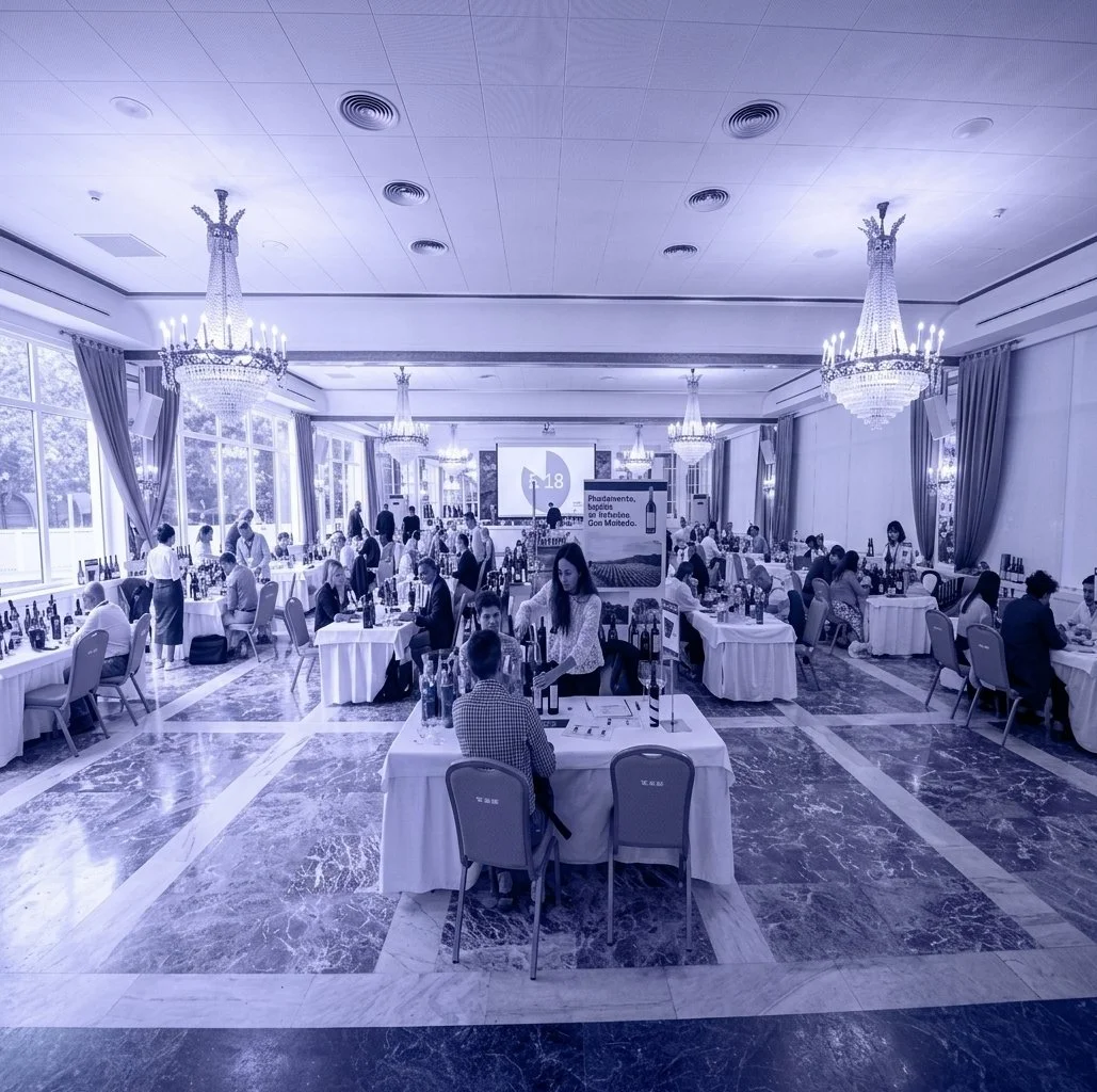Salón de eventos con mesas redondas, personas conversando y decoradas con manteles blancos, con grandes ventanas y candelabros de cristal en el techo.