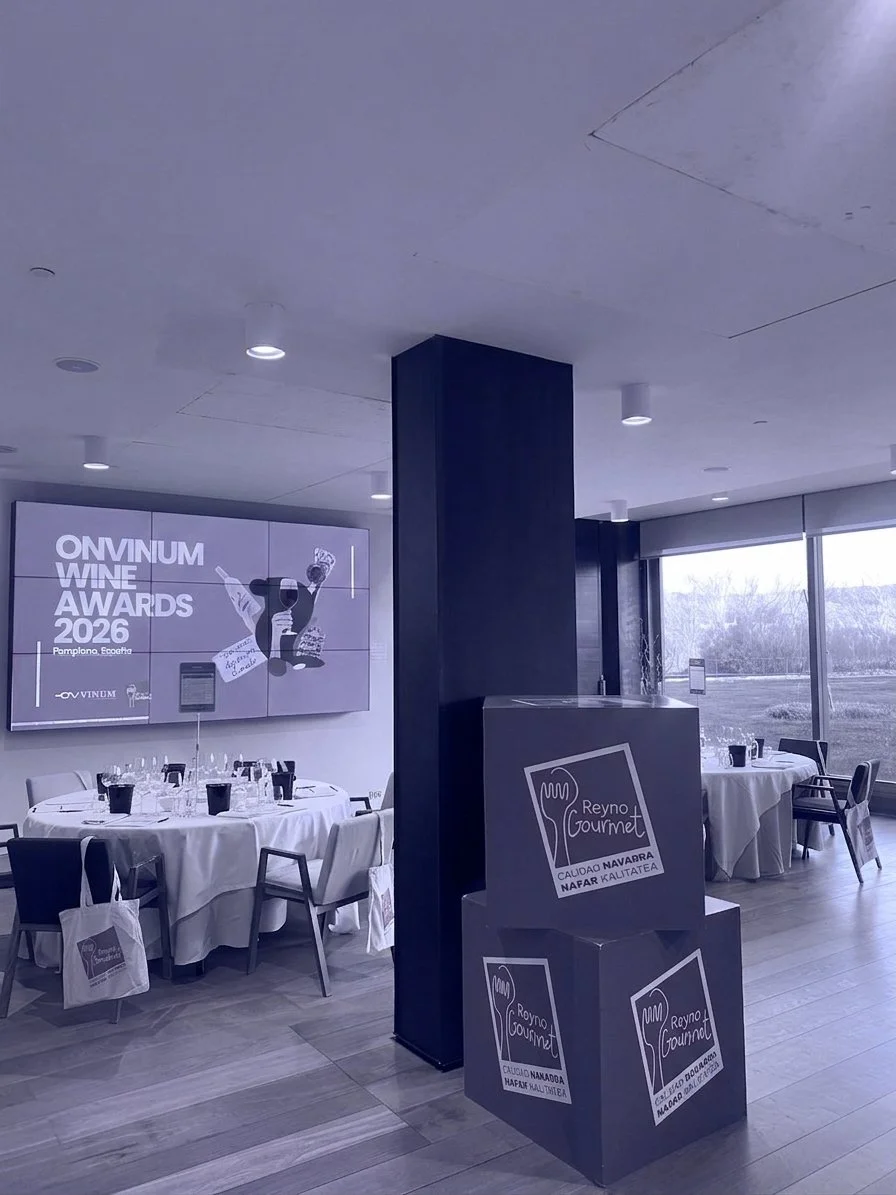 Sala con mesas decoradas para un evento, pantalla con anuncio de premios de vino 2026 y carteles de gastronomía, en un ambiente moderno con ventanas grandes.