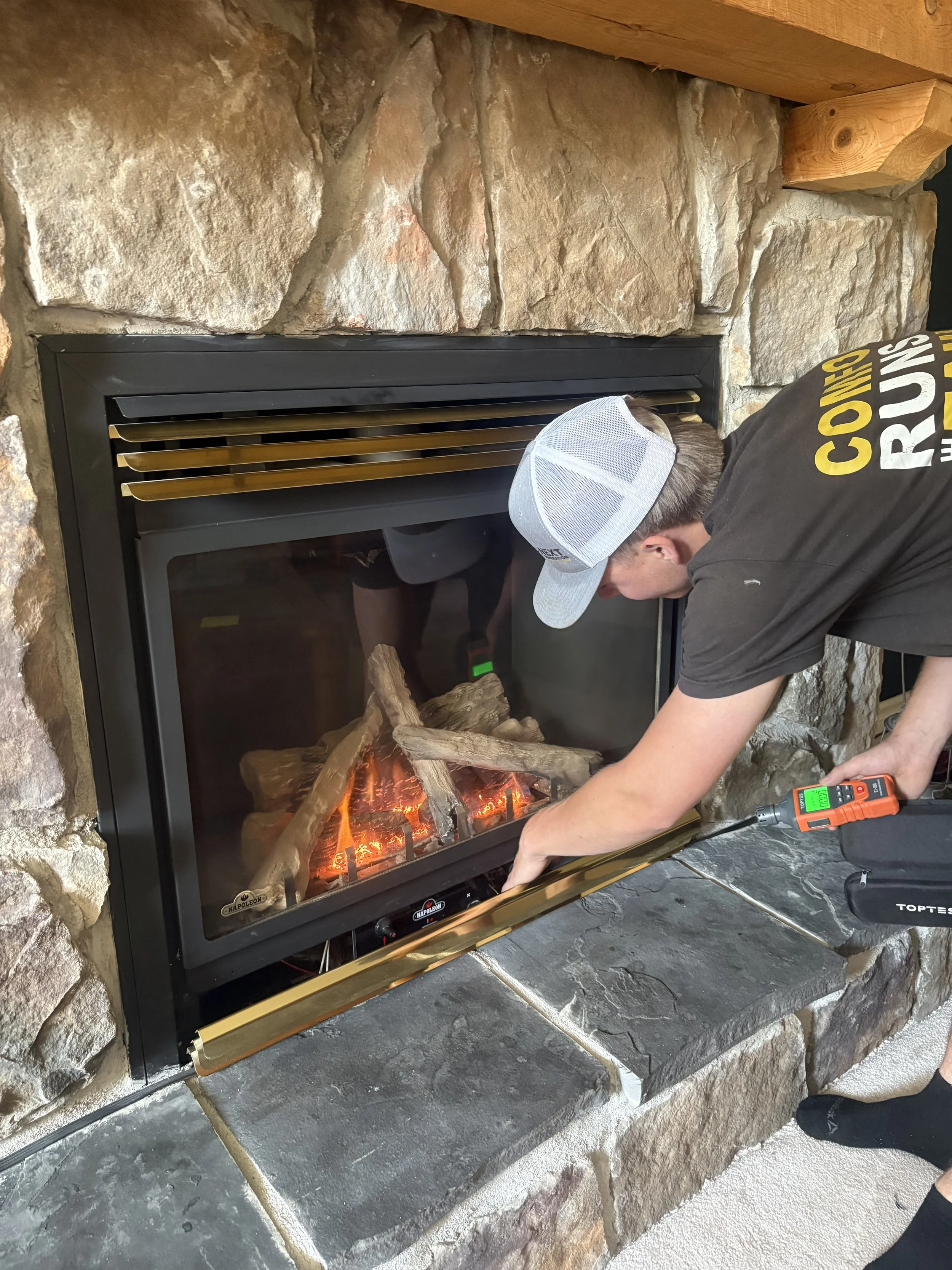 A man with gray hair kneeling in front of a fireplace, working on it with tools, including a drill, wearing a tool belt with various tools, in a living room with a gray couch and green plants.