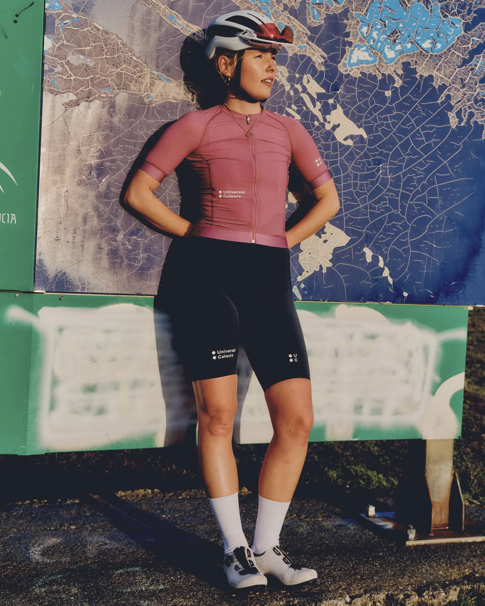 A woman wearing a pink cycling jersey, black shorts, white cycling shoes, white socks, and a helmet, standing against a large map display outdoors.