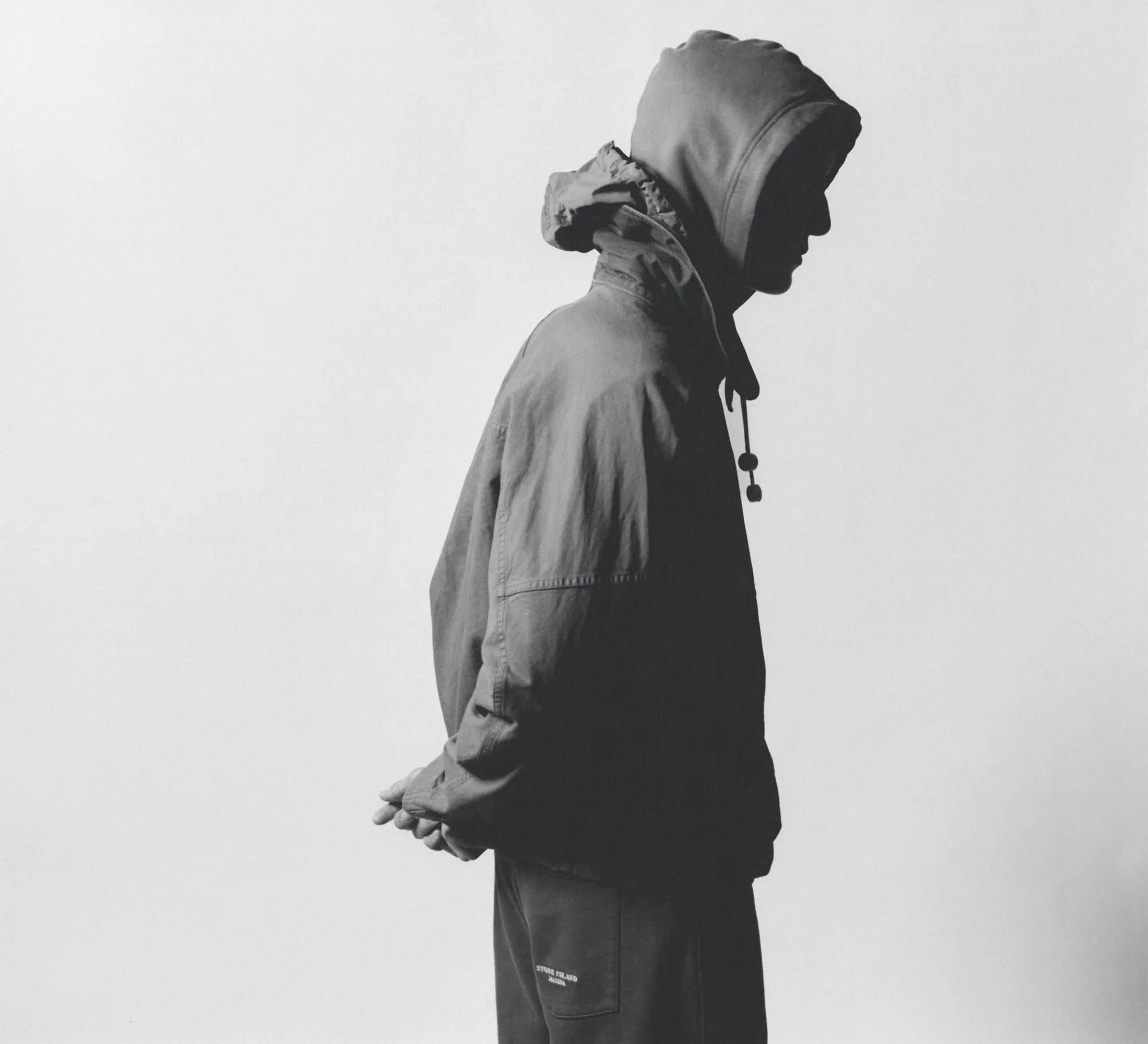 Black and white side profile of a person wearing a hooded jacket and a beanie, with hands clasped in front, against a plain background.