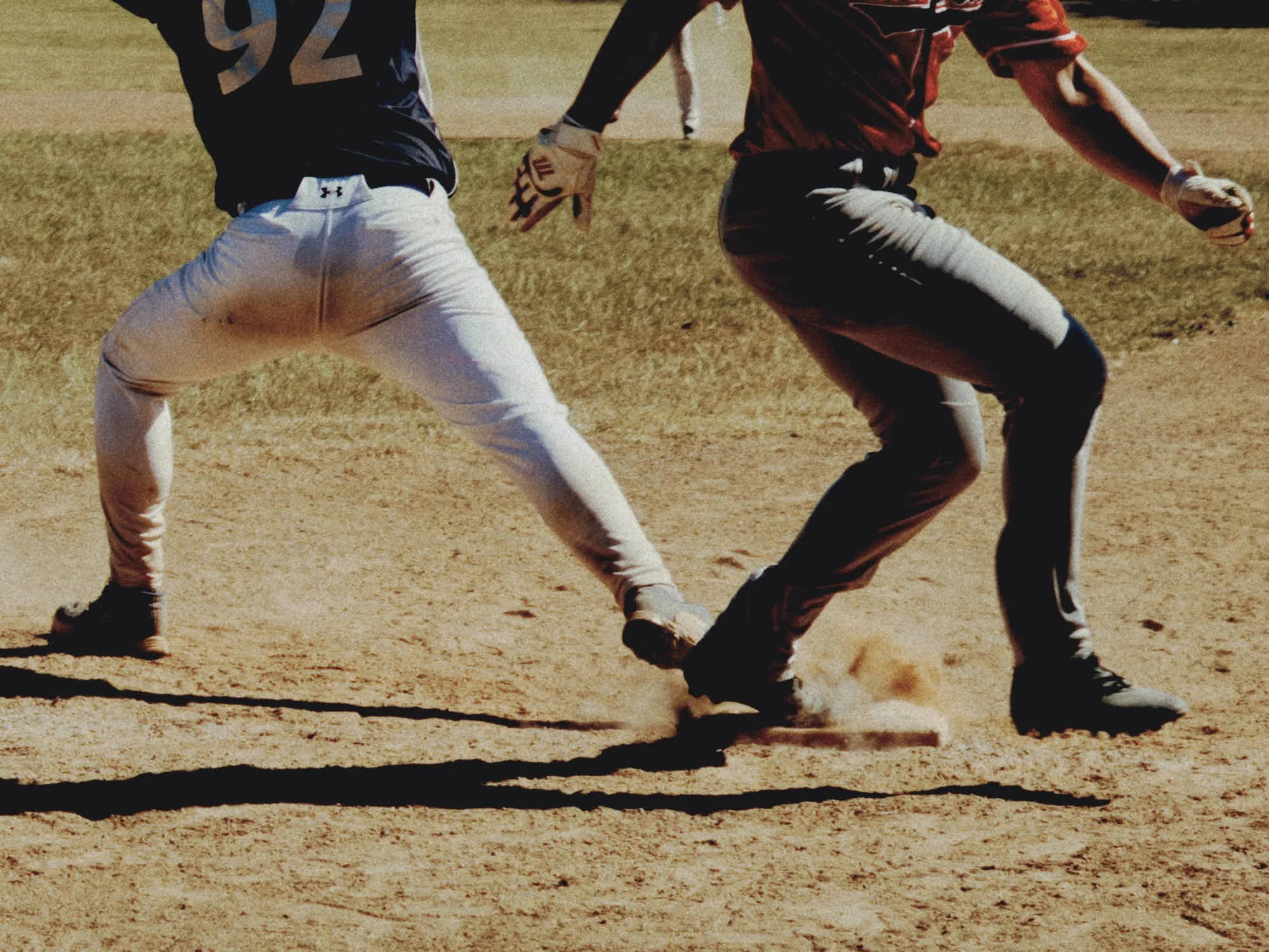 Two baseball players are running on a dirt baseball field, one in a black jersey and white pants, and the other in a red jersey and black pants, with a grassy area in the background.