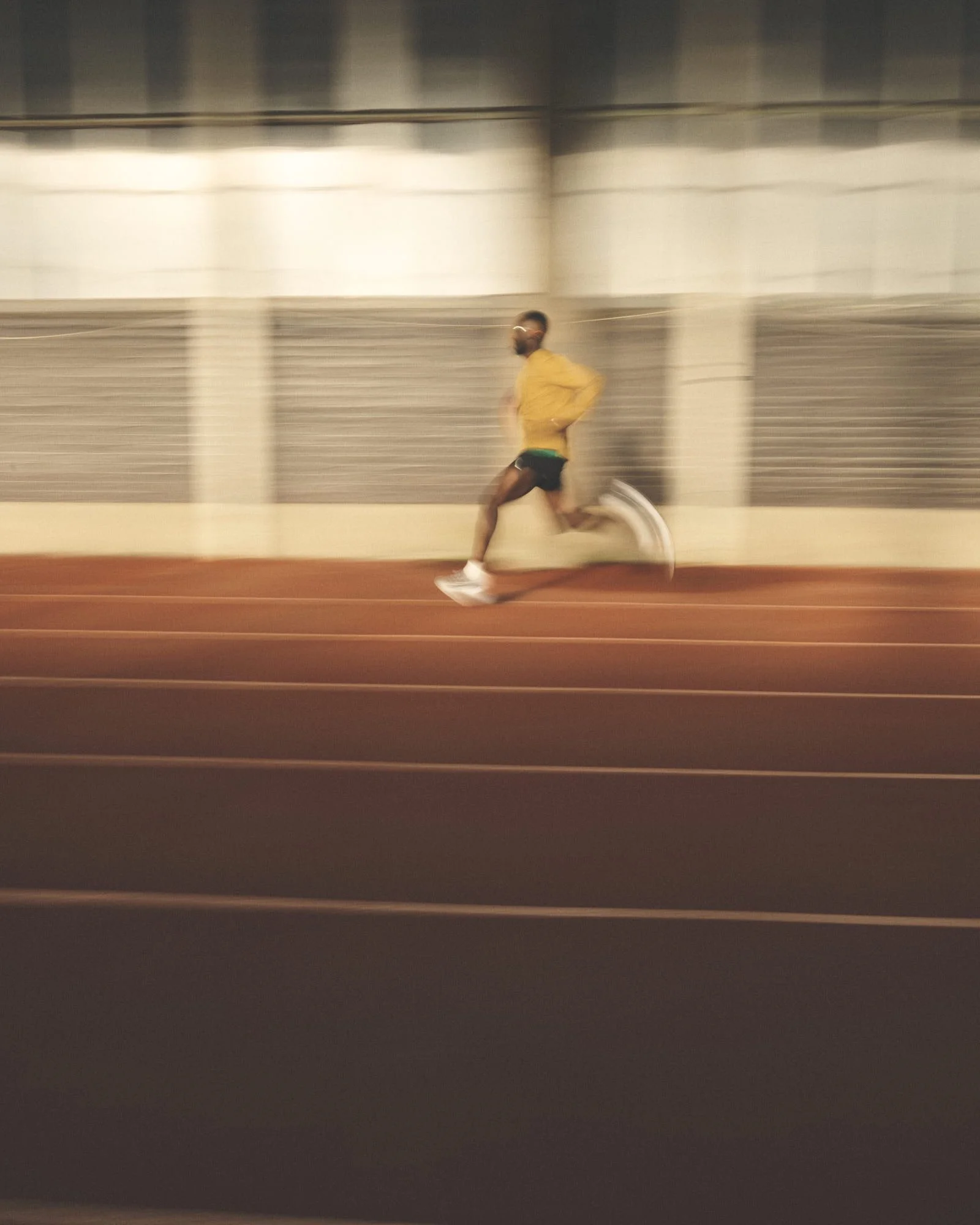 A man jogging on an indoor track with a high ceiling and metal walls, wearing a yellow jacket, black shorts, and white sneakers.