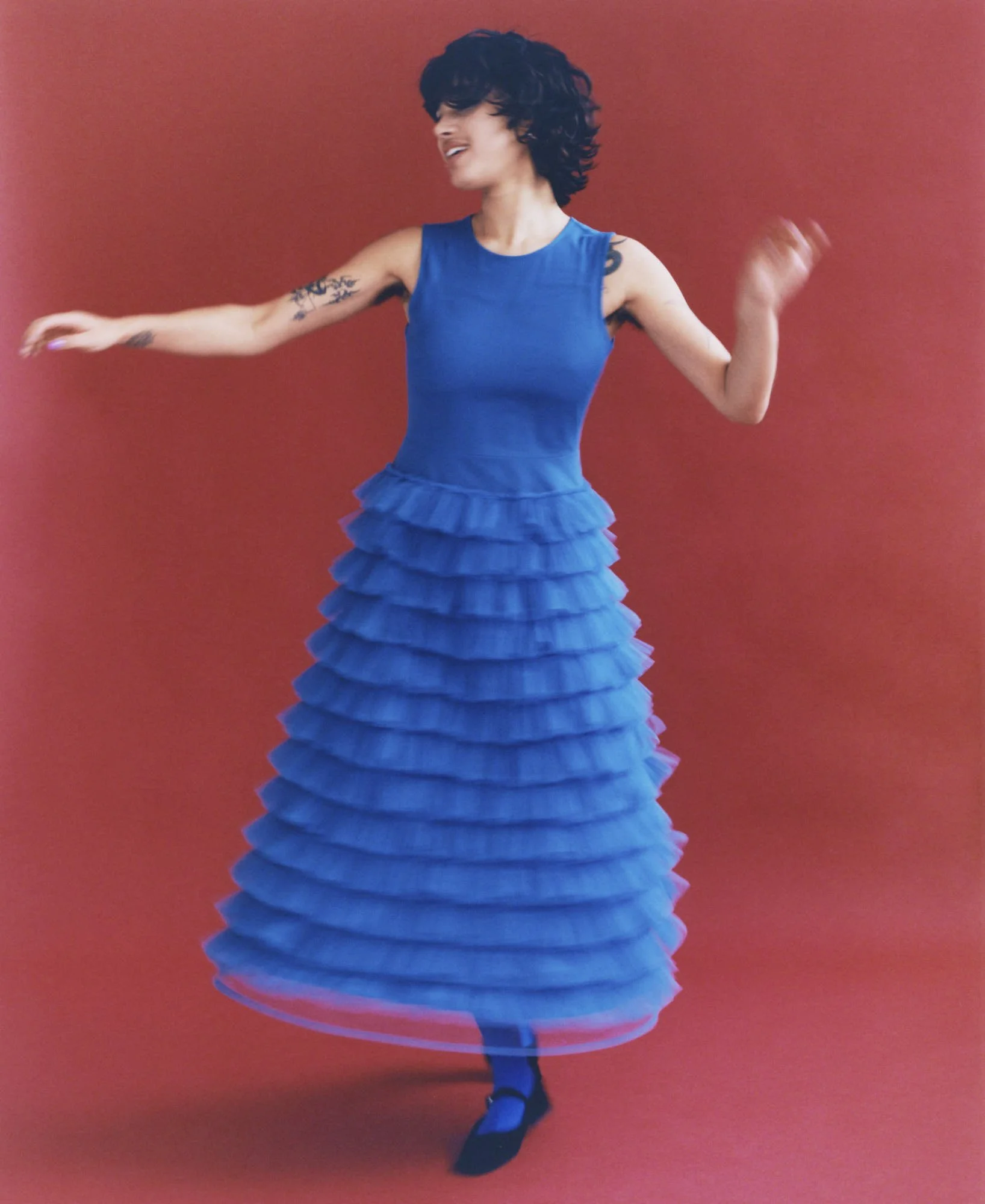 A woman dancing with a red background, wearing a blue sleeveless top and a layered ruffle skirt, captured mid-movement.