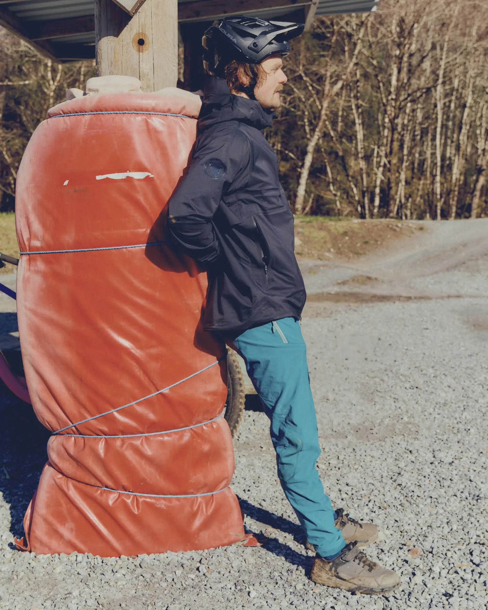 A man wearing a black helmet, black jacket, blue pants, and hiking boots stands next to a large orange bag tied to a wooden post outdoors during daytime. There are trees and a gravel path in the background.