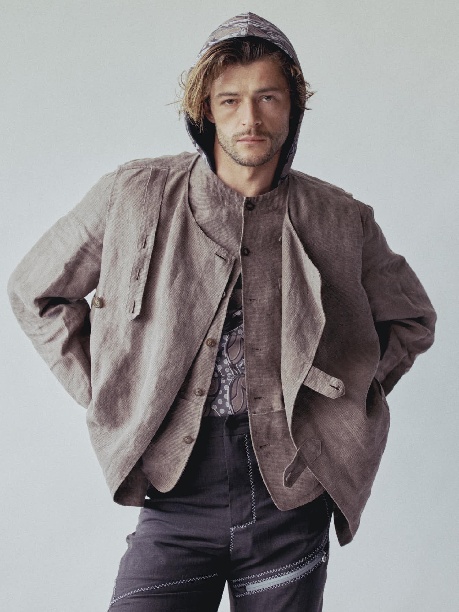 Man wearing a patterned hoodie and a beige jacket, standing against a plain light background, with hands in pockets and a serious expression.