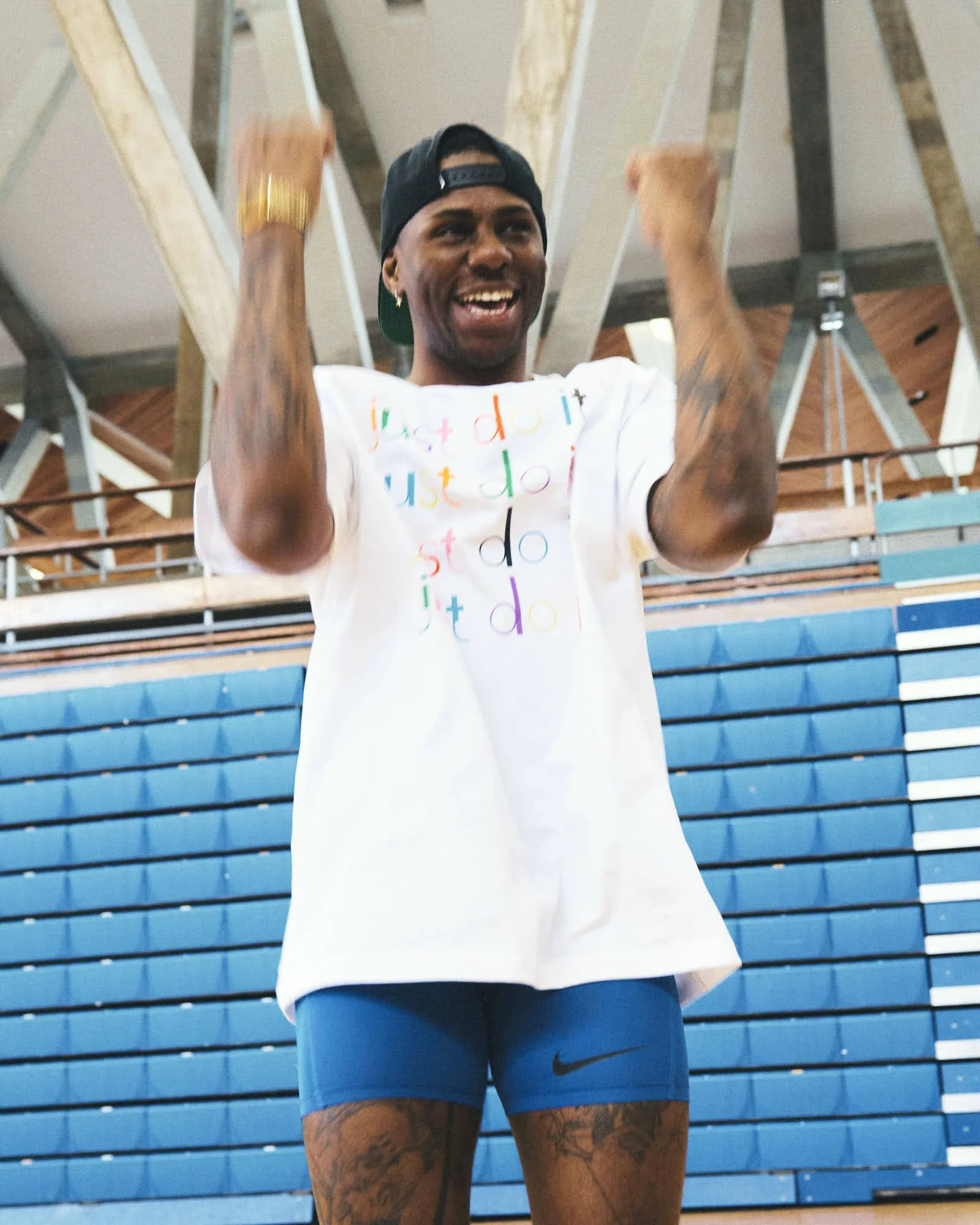A young man celebrating in a gym with blue bleachers in the background. He is wearing a white t-shirt with colorful text, blue Nike shorts, a black cap worn backwards, and a gold watch. He is smiling and raising his fists in excitement.