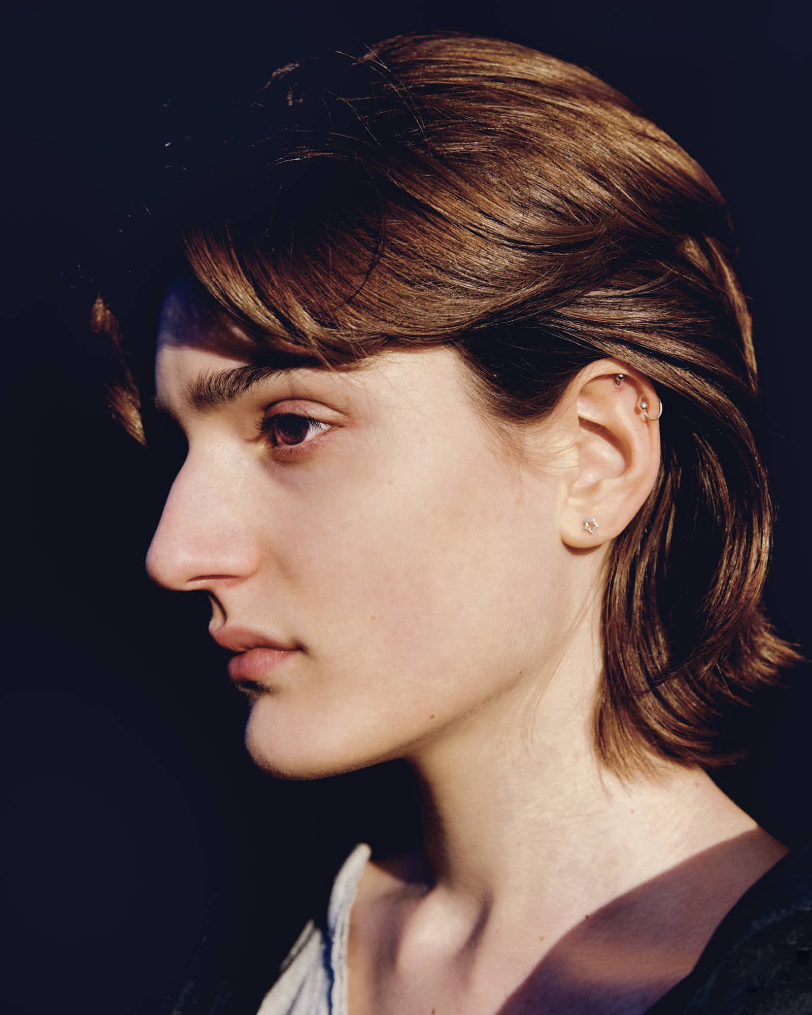A side profile of a woman with short, auburn hair, wearing earrings and a top with a lace trim, against a dark background.