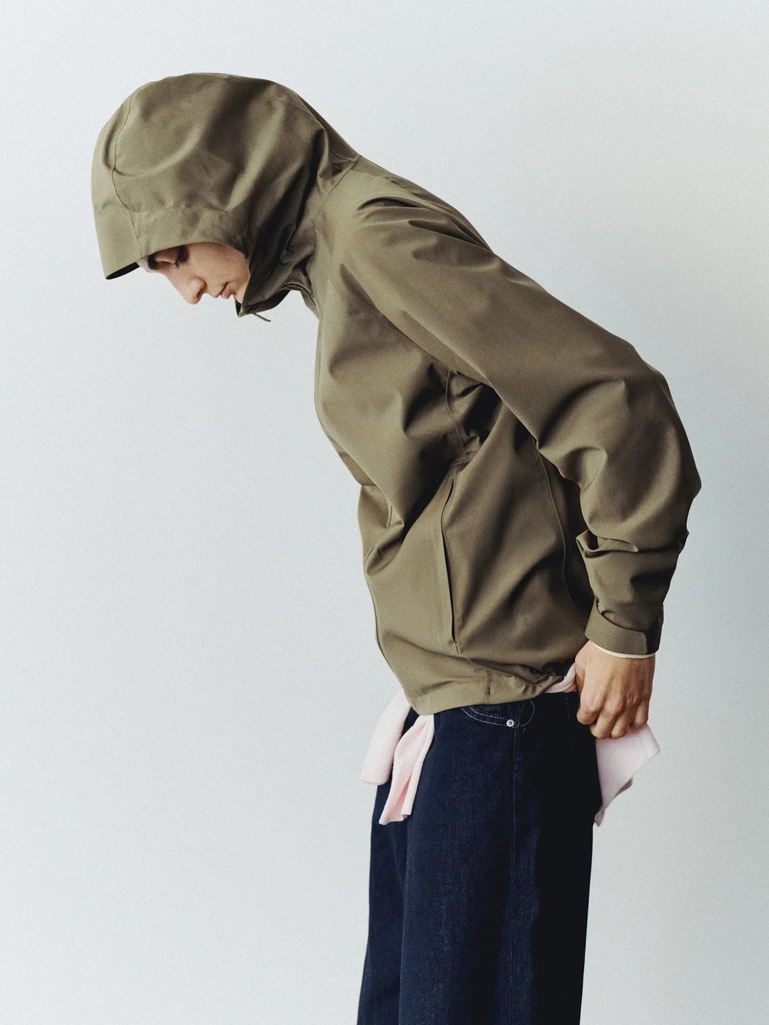A person wearing a khaki hooded raincoat and dark jeans, leaning forward with a pink sweatshirt tied around their waist, against a plain light gray background.