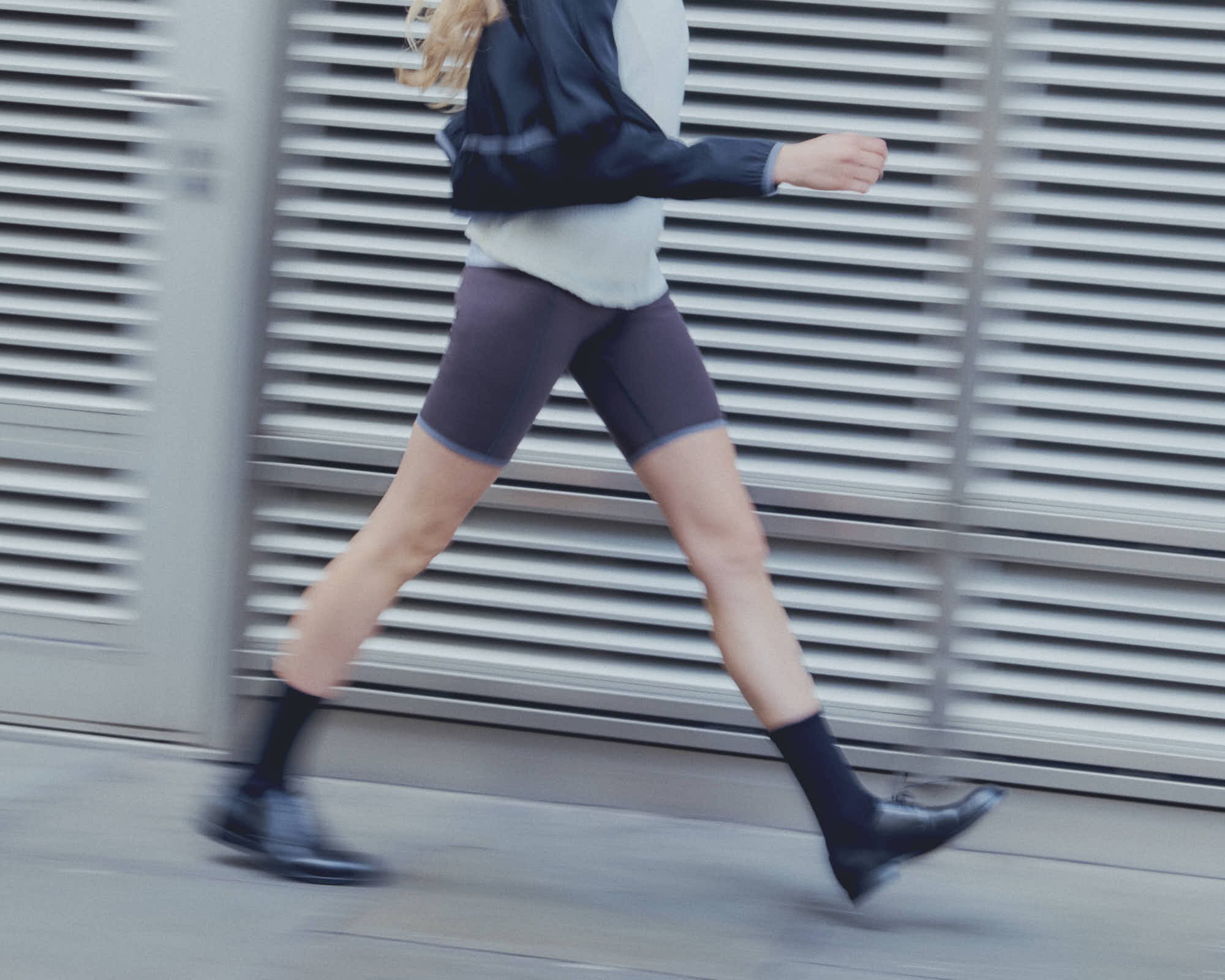 Person with long blond hair running in athletic clothing, including a gray sweatshirt, black shorts, and black shoes, in front of horizontal metallic blinds.