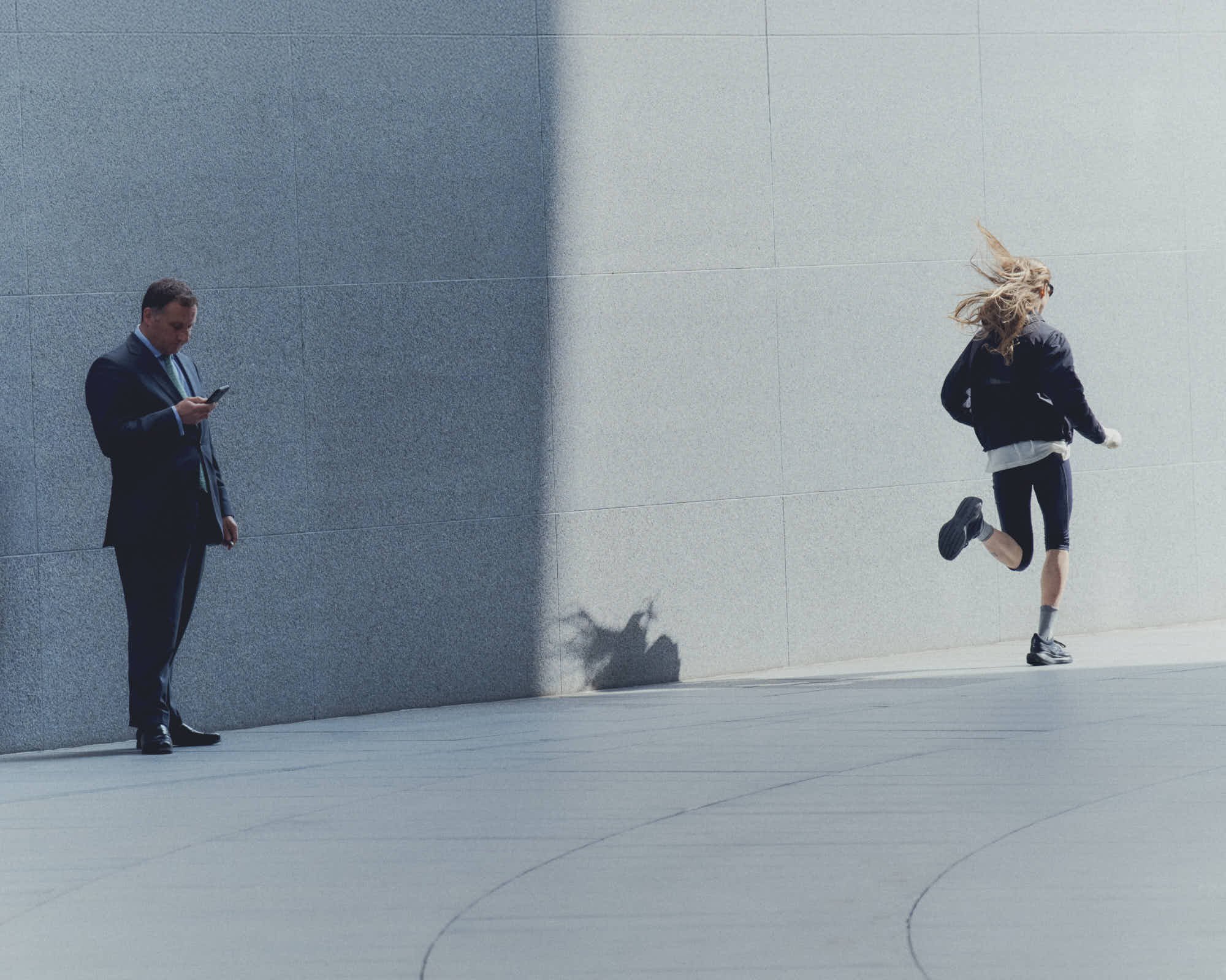 A man in a suit standing against a blue wall, looking at his phone, while a woman with long hair jumping against the same wall, wearing athletic clothing and sneakers.