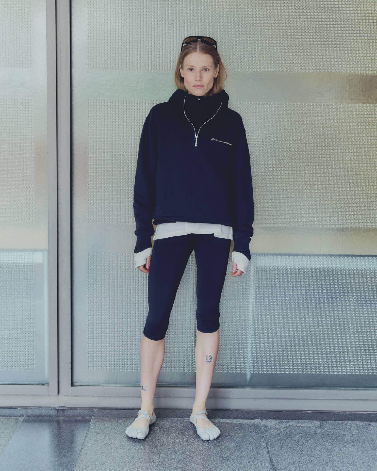 Young woman with short blonde hair wearing black sweatshirt, white T-shirt, black capris, and beige slip-on shoes, standing against a silver perforated metal wall.