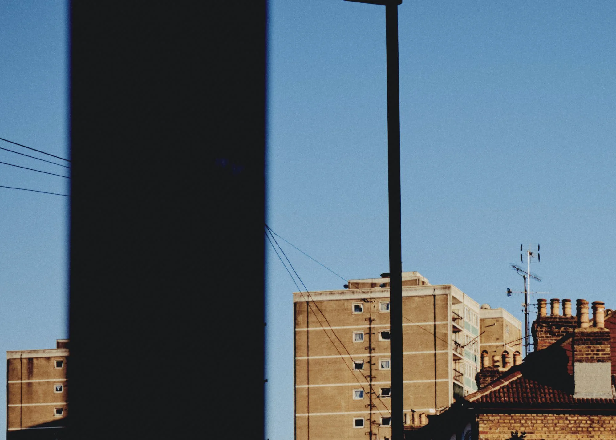 A cityscape with residential buildings, rooftops with chimneys, and an electrical pole with wires against a clear blue sky, reflected in a glass window that creates a distorted view.