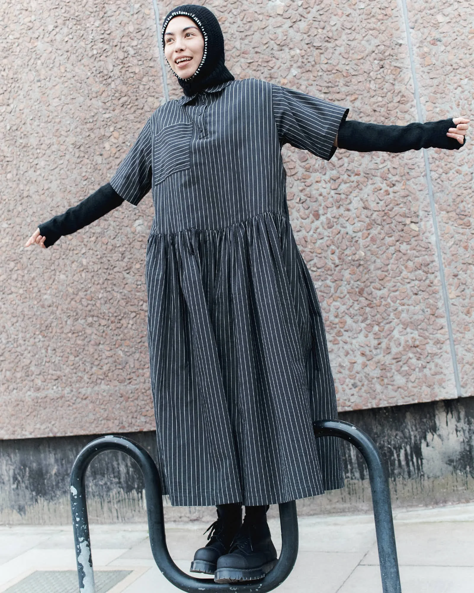 A woman in a black hoodie and black platform shoes standing on a circular metal structure outdoors against a textured beige wall.