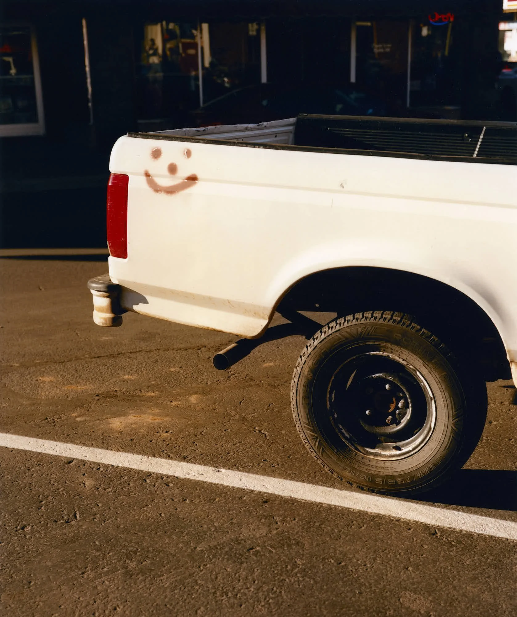 A white pickup truck with a smiley face drawn on the side near the rear. The truck is parked in a parking lot with a marked parking space visible on the ground. The background is dark and shows some storefronts and a red neon sign.