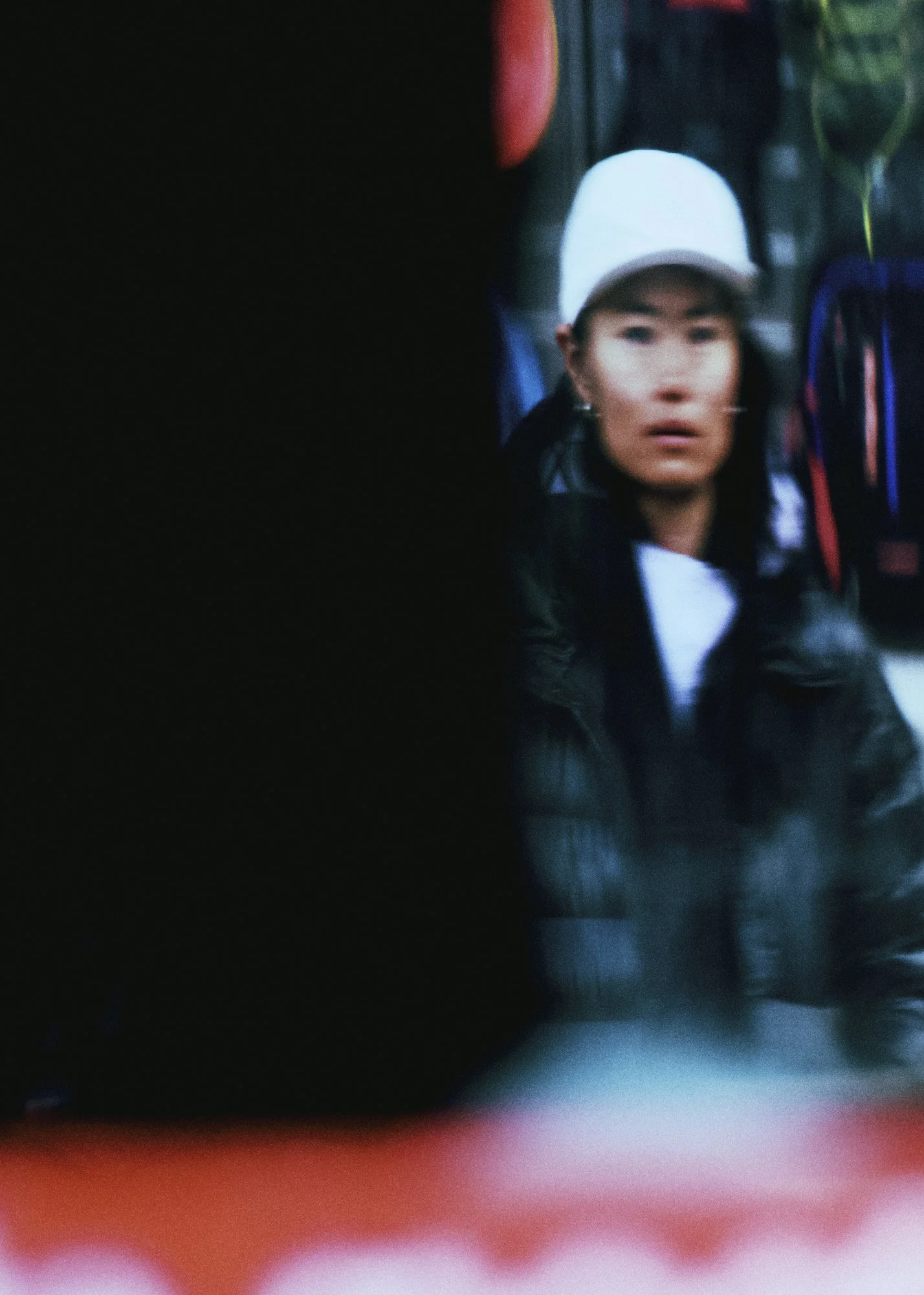 Blurred close-up shot of a woman with a white cap and dark clothing, with colorful objects and clothing surrounding her in the background.