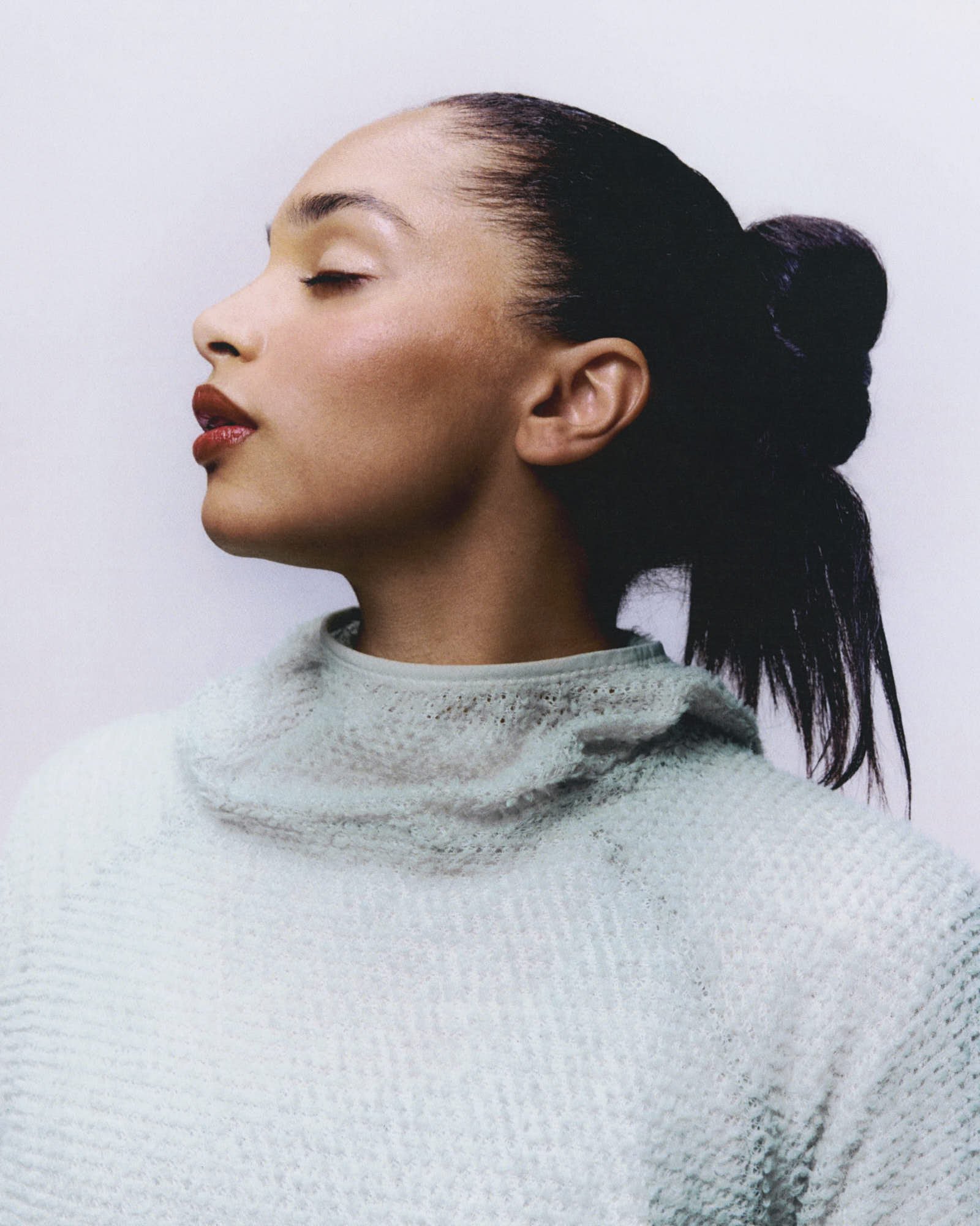 Close-up side profile of a woman with her eyes closed, wearing red lipstick and a light-colored sweater, with dark hair styled in a sleek bun.