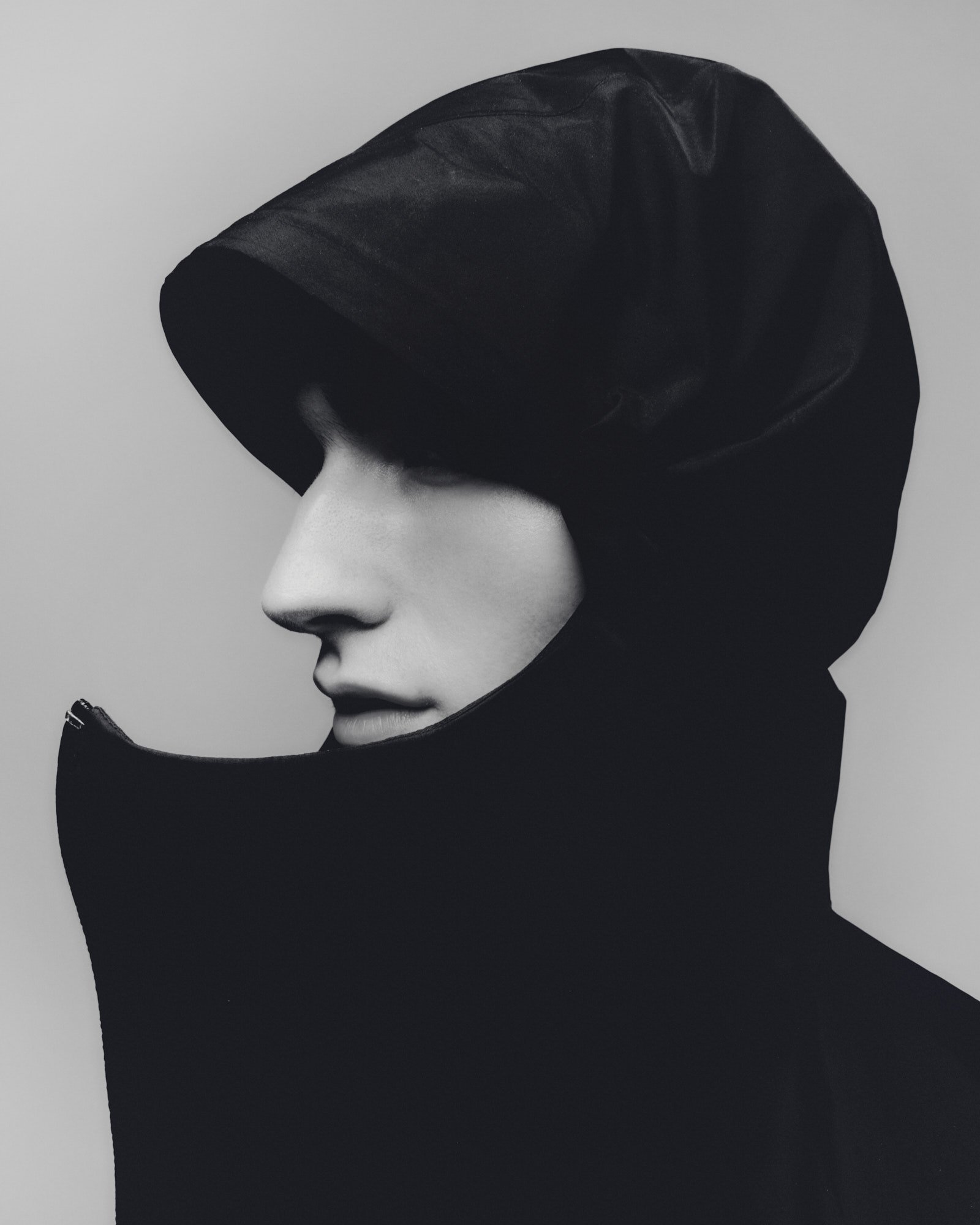 A black mannequin head wearing a black velvet or satin hooded garment against a plain light gray background.