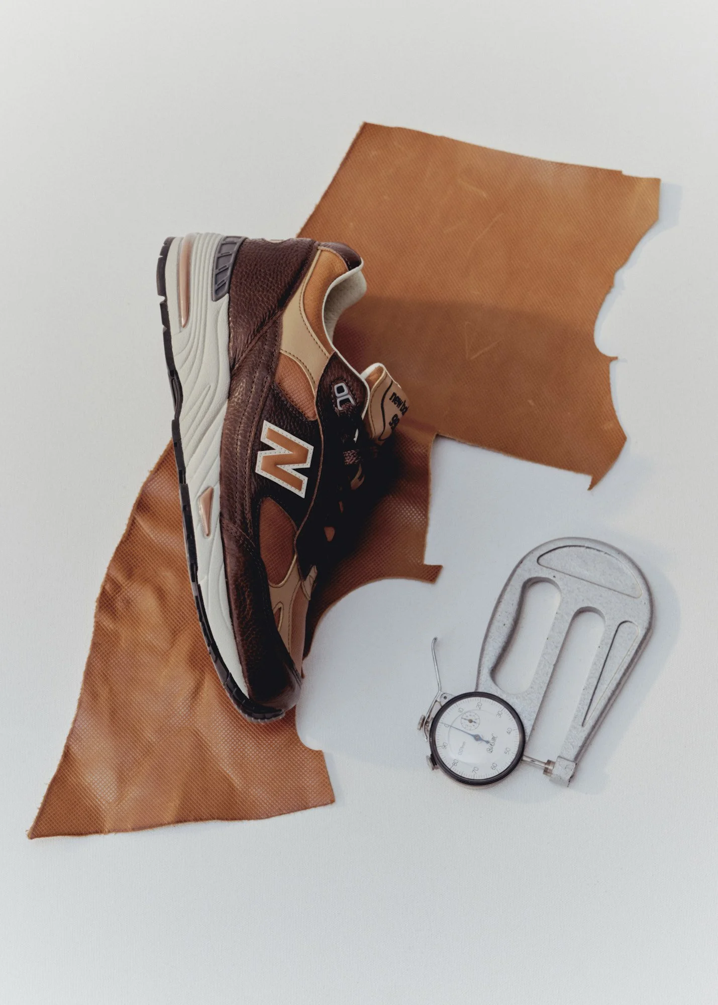 A pair of sneakers with brown, beige, and orange colors resting on an orange cloth, a mechanical watch, a sewing gauge, and a small round stopwatch on a white surface.