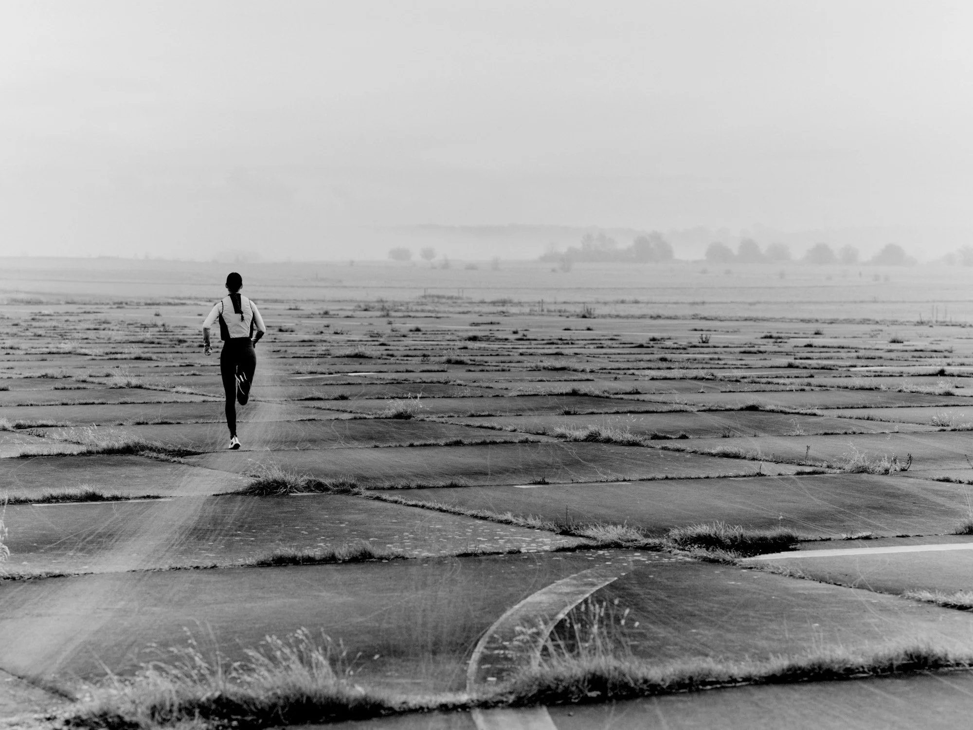 A person walking away in a vast, empty parking lot. The scene is in black and white, with overcast sky and distant trees.