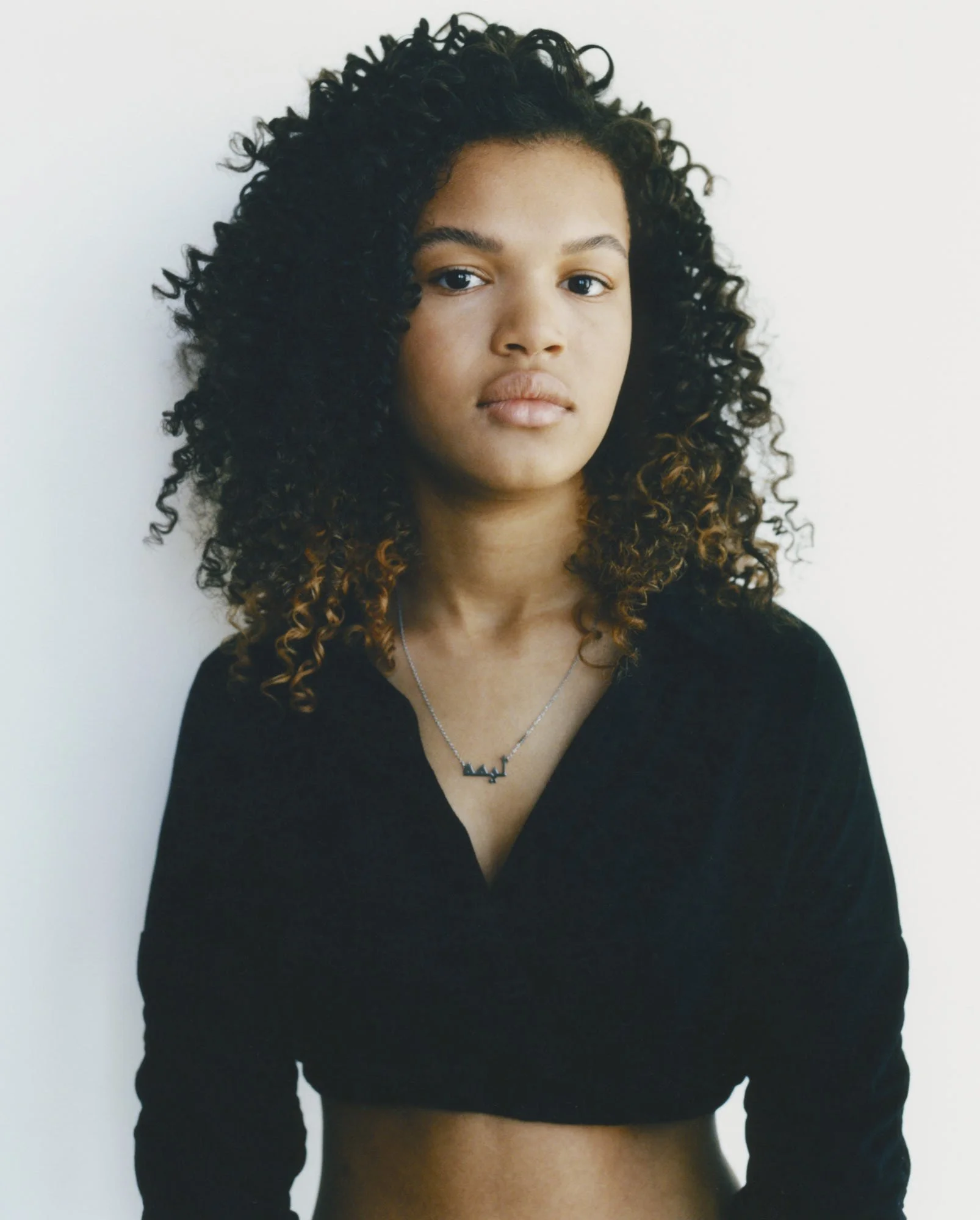 A woman with dark, curly hair with caramel tips, wearing a black V-neck top and a silver necklace with a mountain-shaped pendant, standing against a white background.