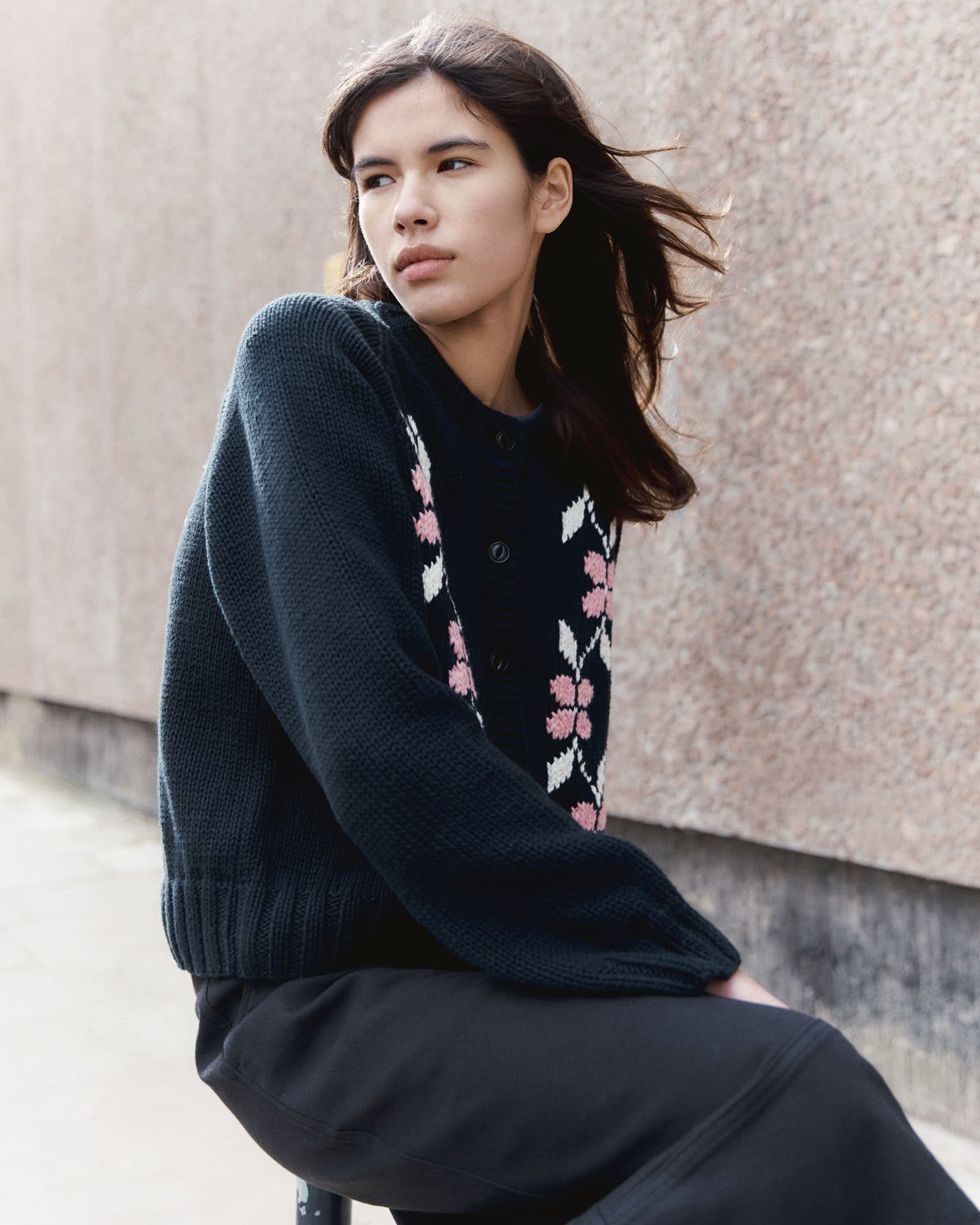 A young woman with dark brown hair sitting against a beige stone wall, wearing a navy knitted cardigan with pink and white patterns and black pants.