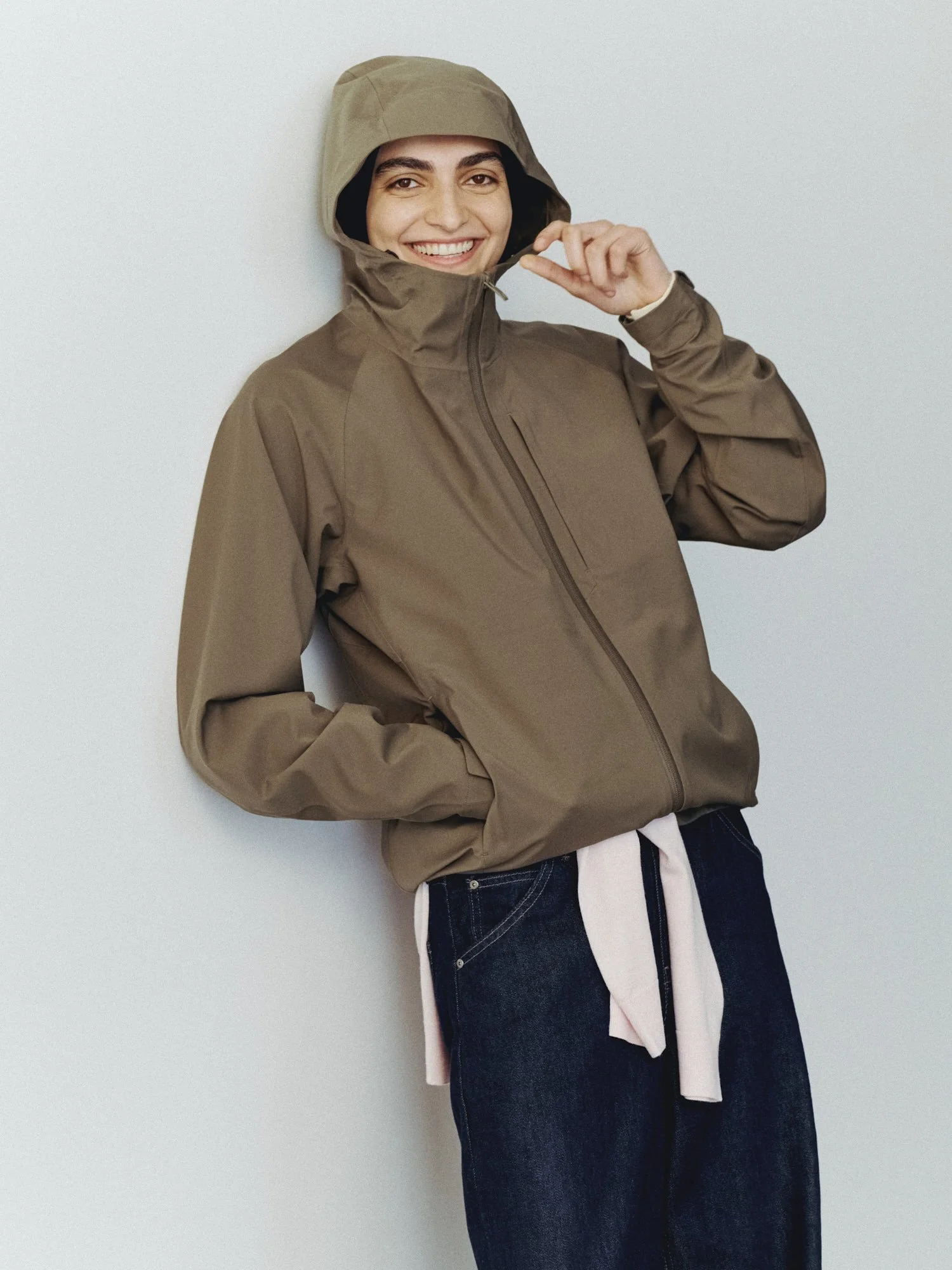 A woman wearing a tan jacket and dark jeans with a pink shirt underneath, smiling and adjusting her jacket's hood, standing against a plain, light-colored wall.