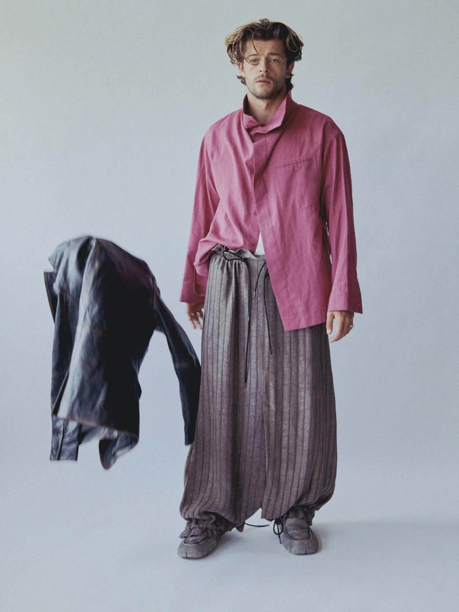 A man with wavy hair and a scruffy face wears a pink satin shirt with an open collar and untucked striped wide-leg pants. He stands barefoot on a plain grey background, looking at the camera with a relaxed expression.