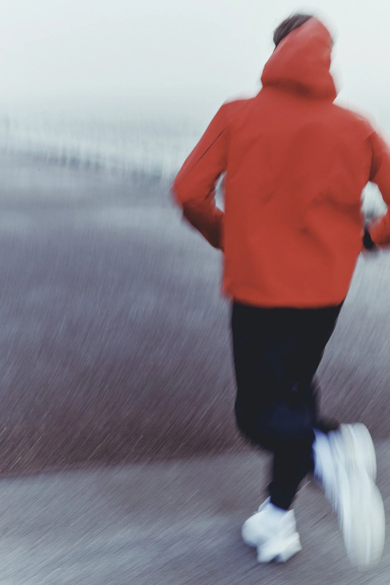 A person running outdoors on a foggy day. The person is wearing a bright orange jacket with the hood up, dark pants, and white sneakers.