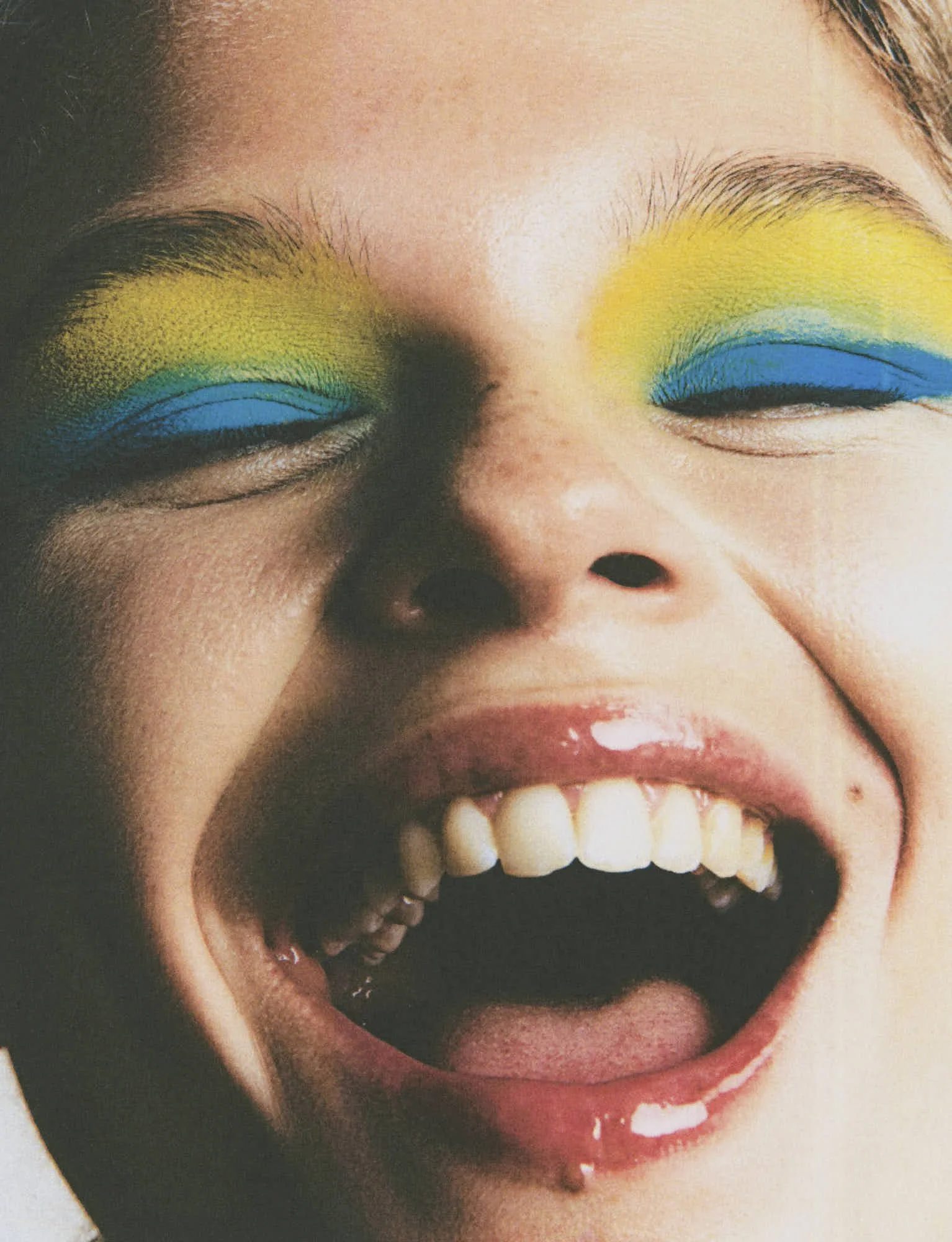 Close-up of a woman's face with vibrant yellow and blue eyeshadow, smiling with visible teeth.
