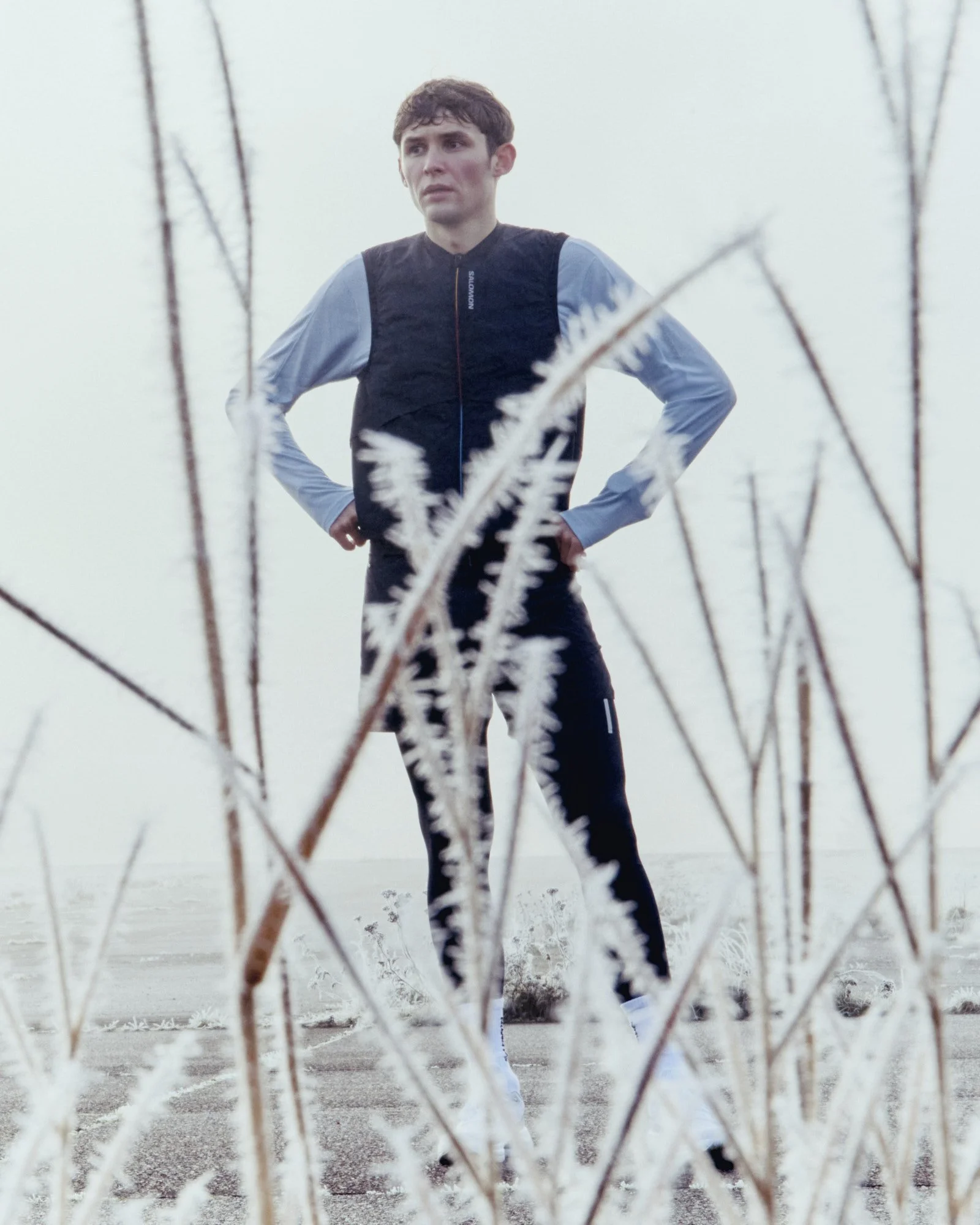 A young man in athletic clothing stands with hands on hips, framed by tall, frosted dried plants on a snowy, overcast outdoor court.