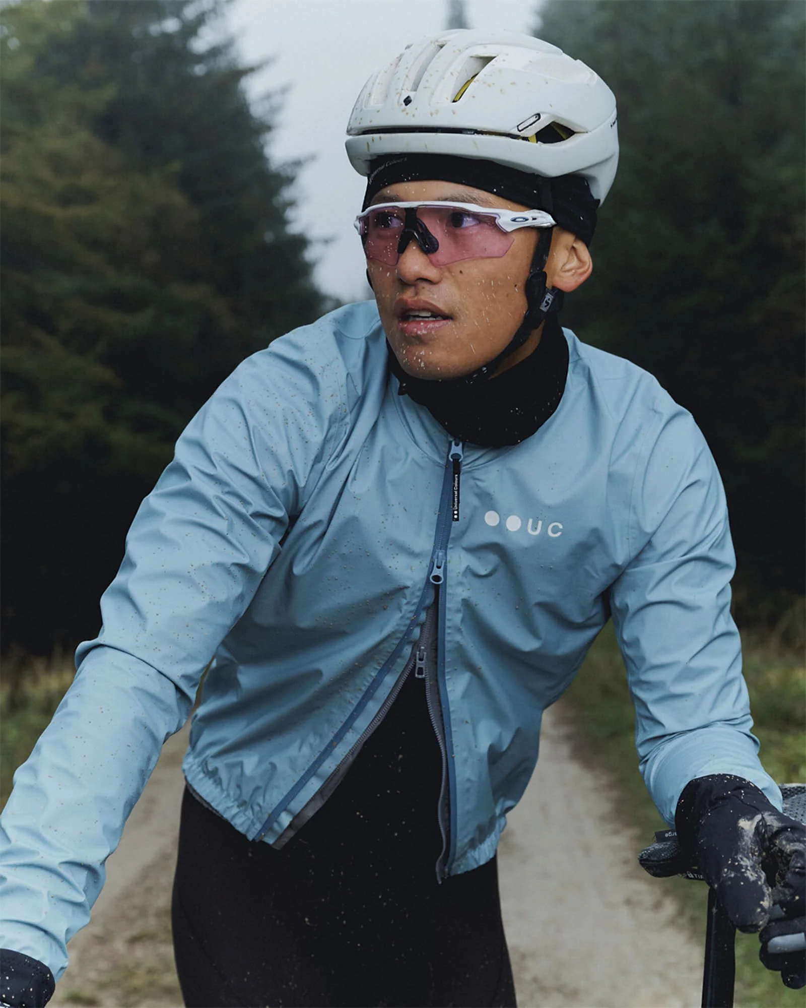 A cyclist wearing a white helmet, pink sunglasses, and a light blue jacket riding outdoors on a dirt path surrounded by green trees, with water splashes visible, indicating wet conditions.
